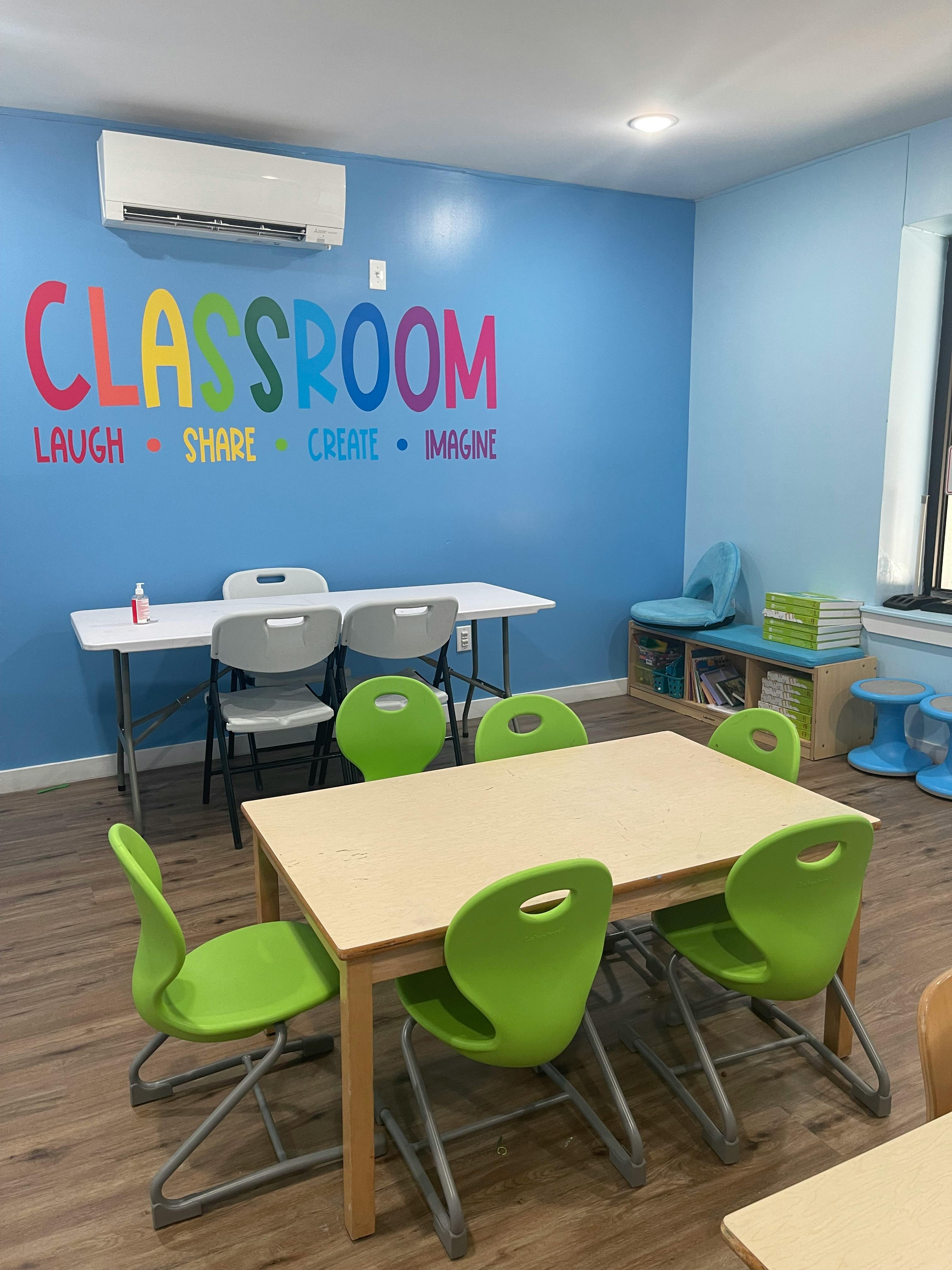 Classroom with tables and chairs, text on wall says "CLASSROOM LAUGH SHARE CREATE IMAGINE"
