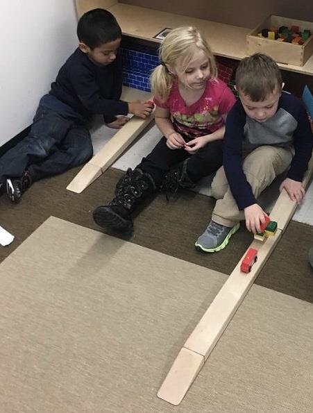 Three children playing with wooden ramps and toy cars on the floor