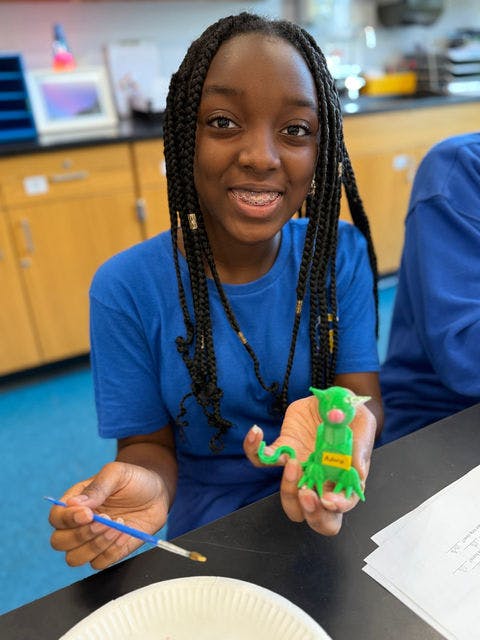 Smiling teen holding a green clay sculpture and a paintbrush in a classroom