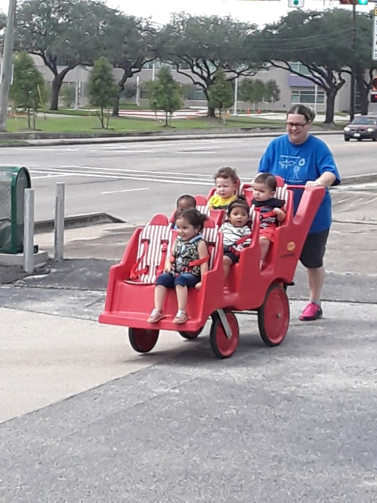 Person pushing multiple children in a red multi-seat stroller on a sidewalk