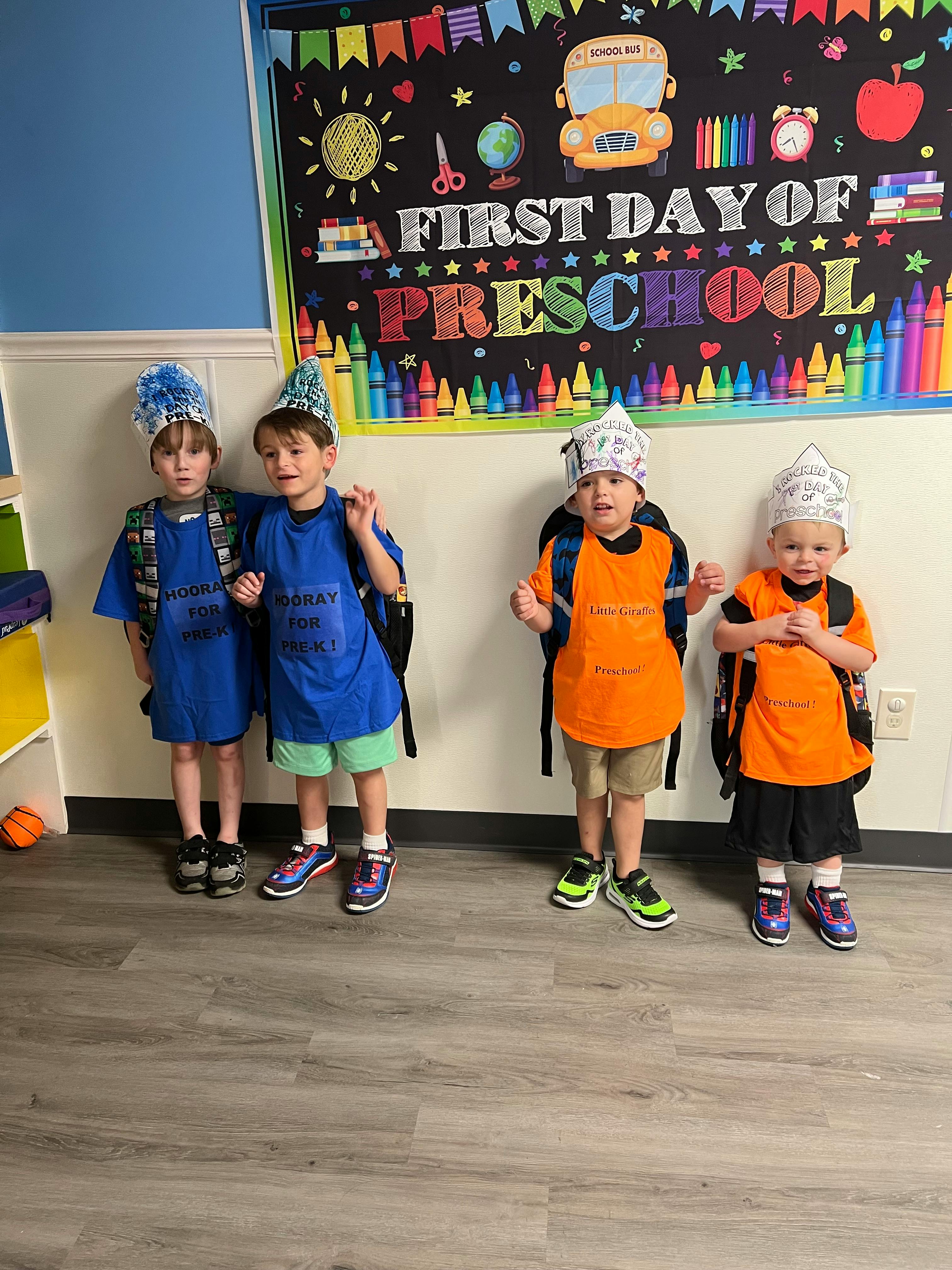 Four children wearing party hats and backpacks in front of a "First Day of Preschool" sign