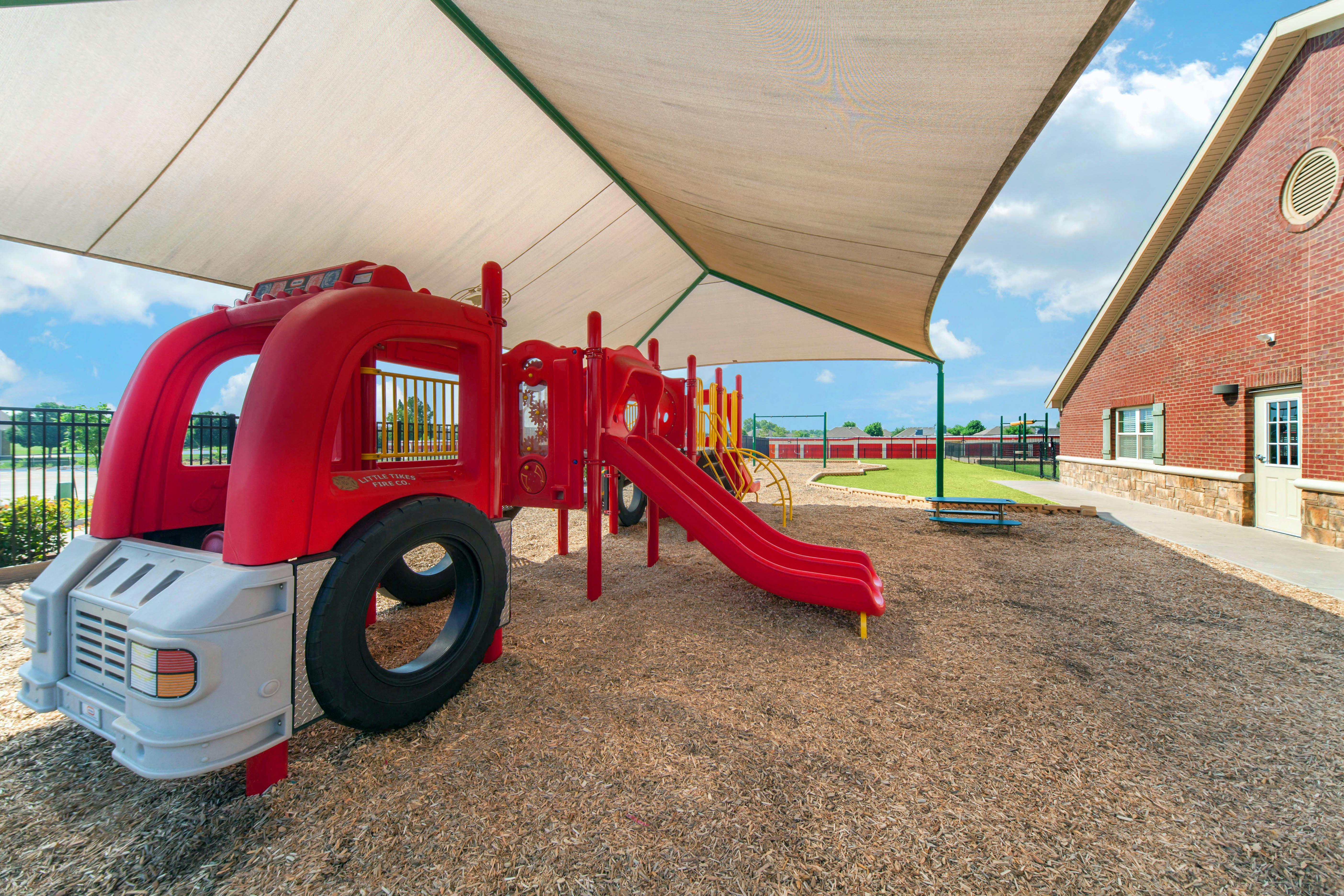 Red playground fire truck structure with slides on wood chips under a shaded canopy