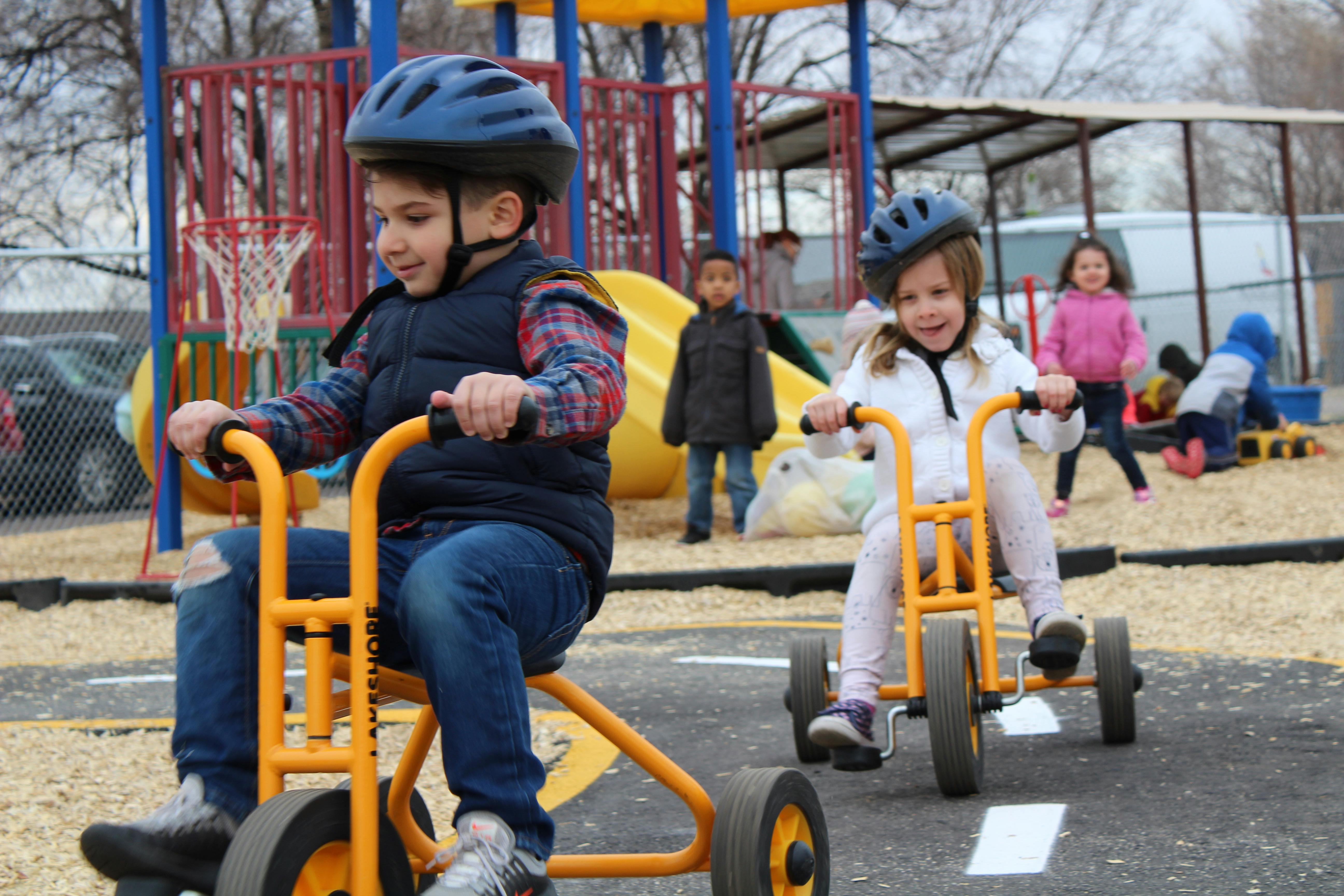 Two children wearing helmets riding tricycles on a playground