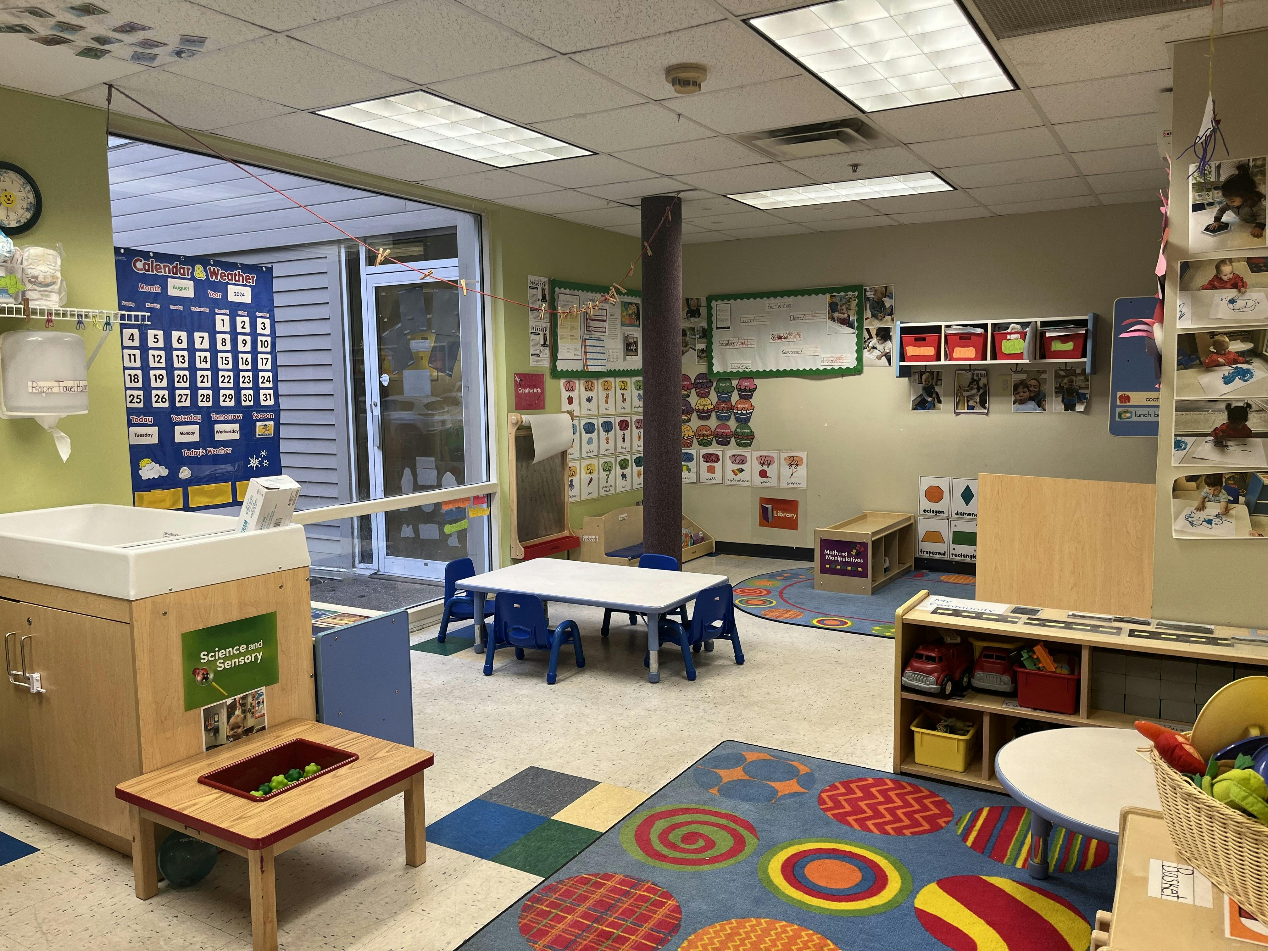 Classroom with child-sized tables and chairs, a changing station, and learning charts on the walls