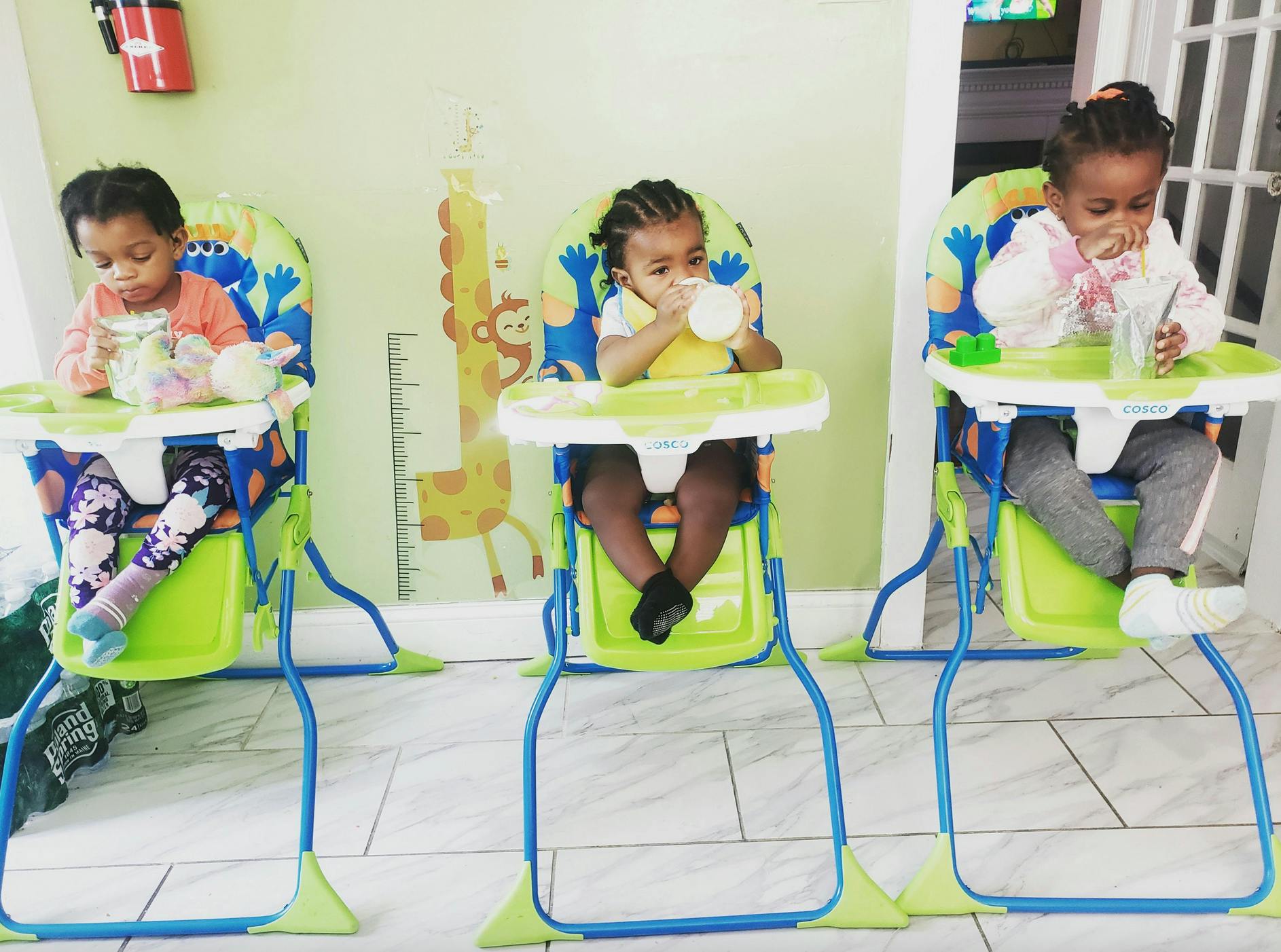 Three children sitting in high chairs drinking from bottles