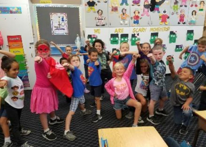 Group of children and an adult posing like superheroes in a classroom