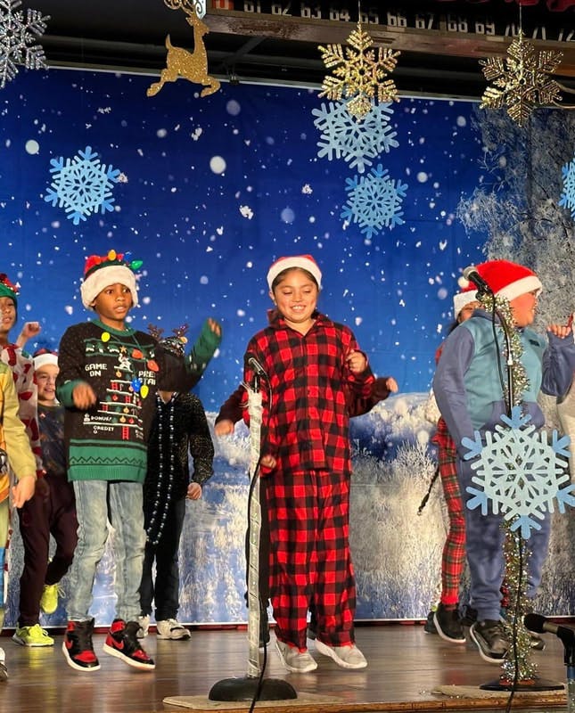 Children wearing Santa hats and festive clothes on a stage with snowflake decorations