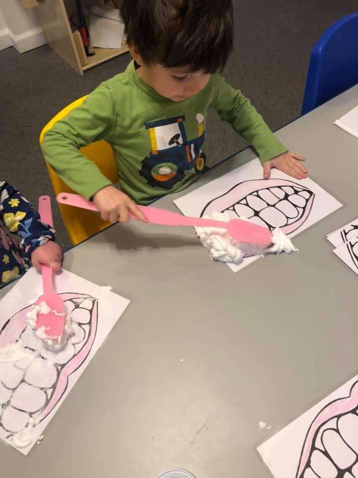 Child using a pink spatula to spread white foam on a drawing of teeth
