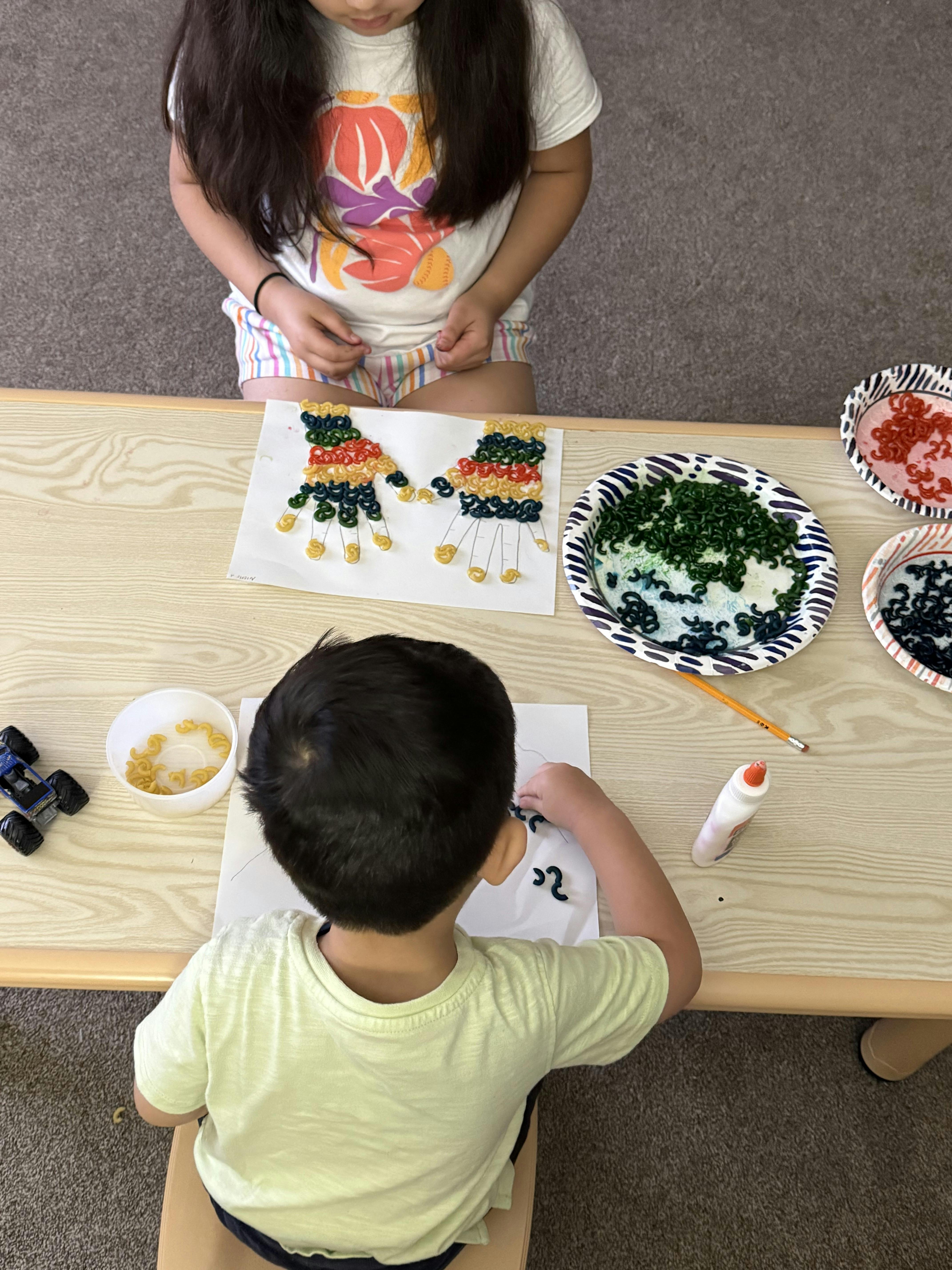 Two children making craft art with glued pasta on paper at a table