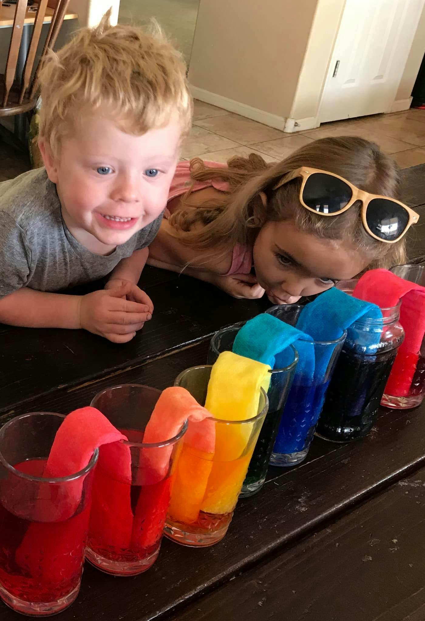 Two children looking at glasses with colored liquid and fabric strips