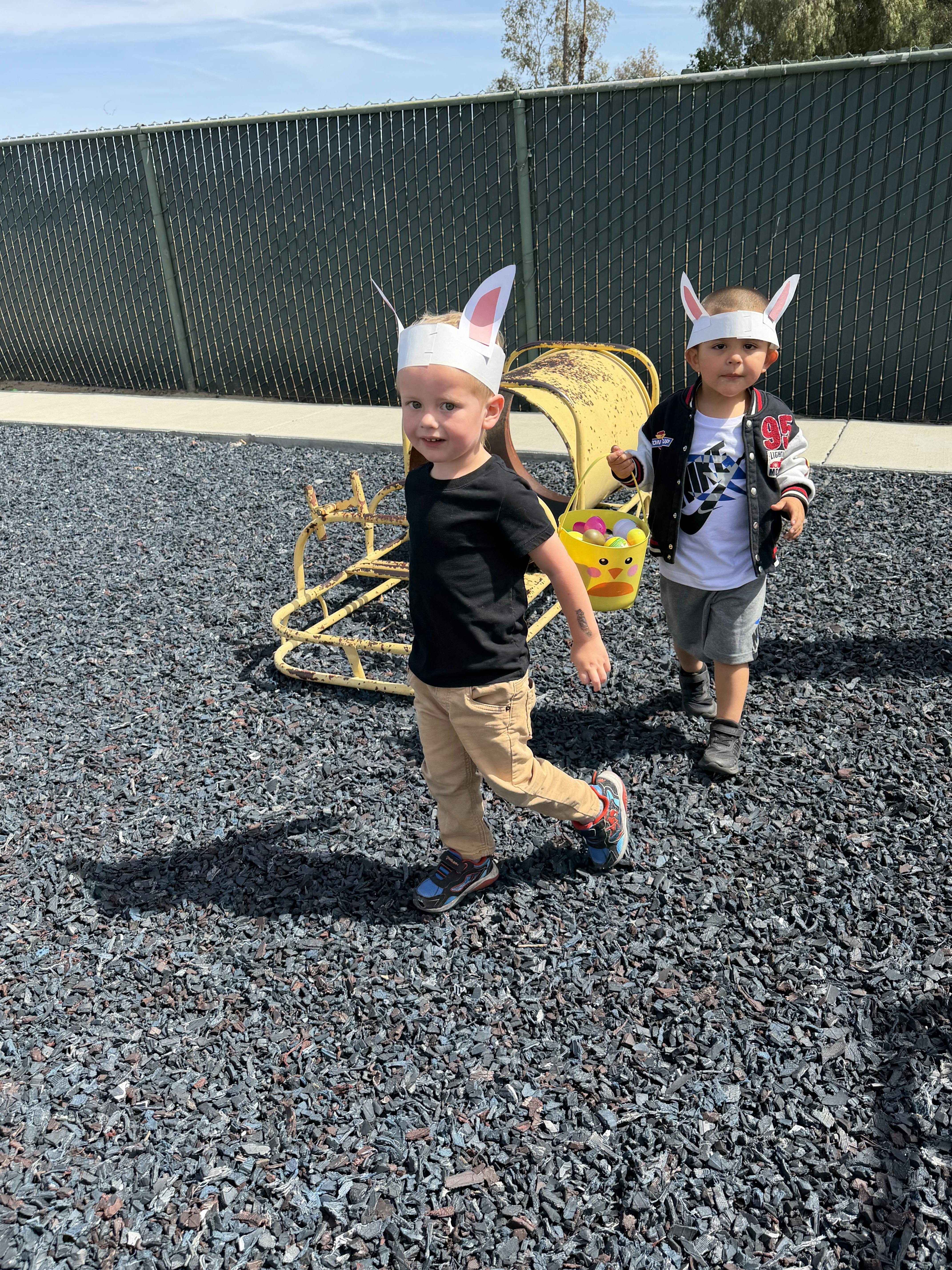 Two children wearing bunny ears at a playground with an Easter basket