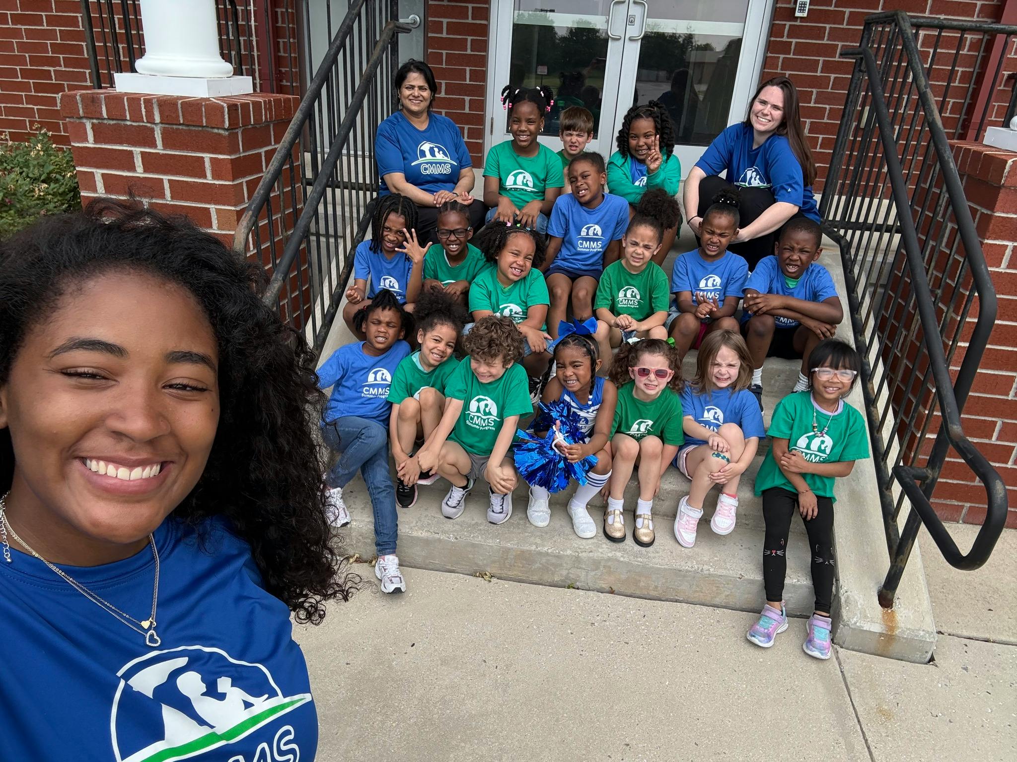 A person taking a selfie with two adults and a group of children on steps