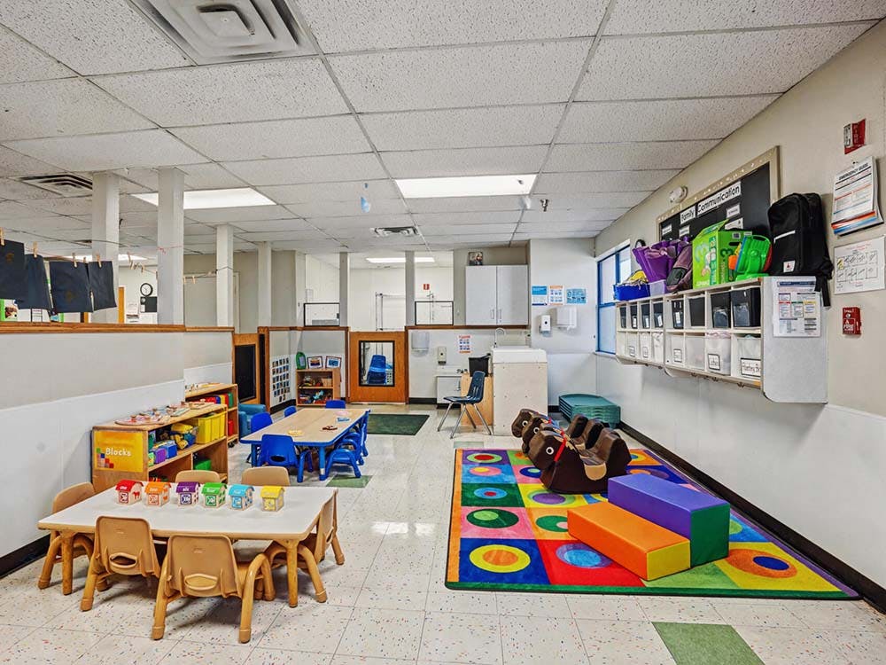 An empty childcare classroom with small tables and chairs, and a colorful rug with play items