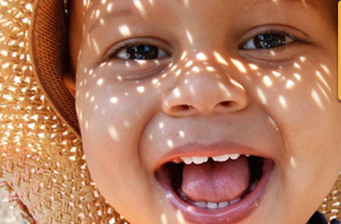 Child wearing a straw hat with dappled sunlight on face, mouth open