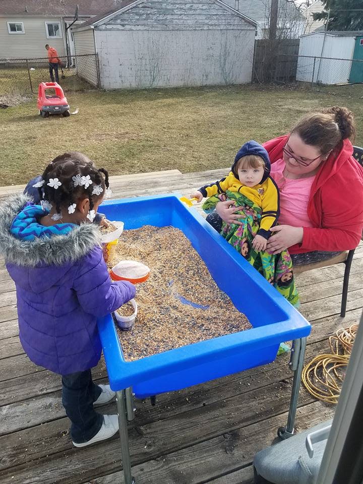 Person holding a child while another child plays at a sand table