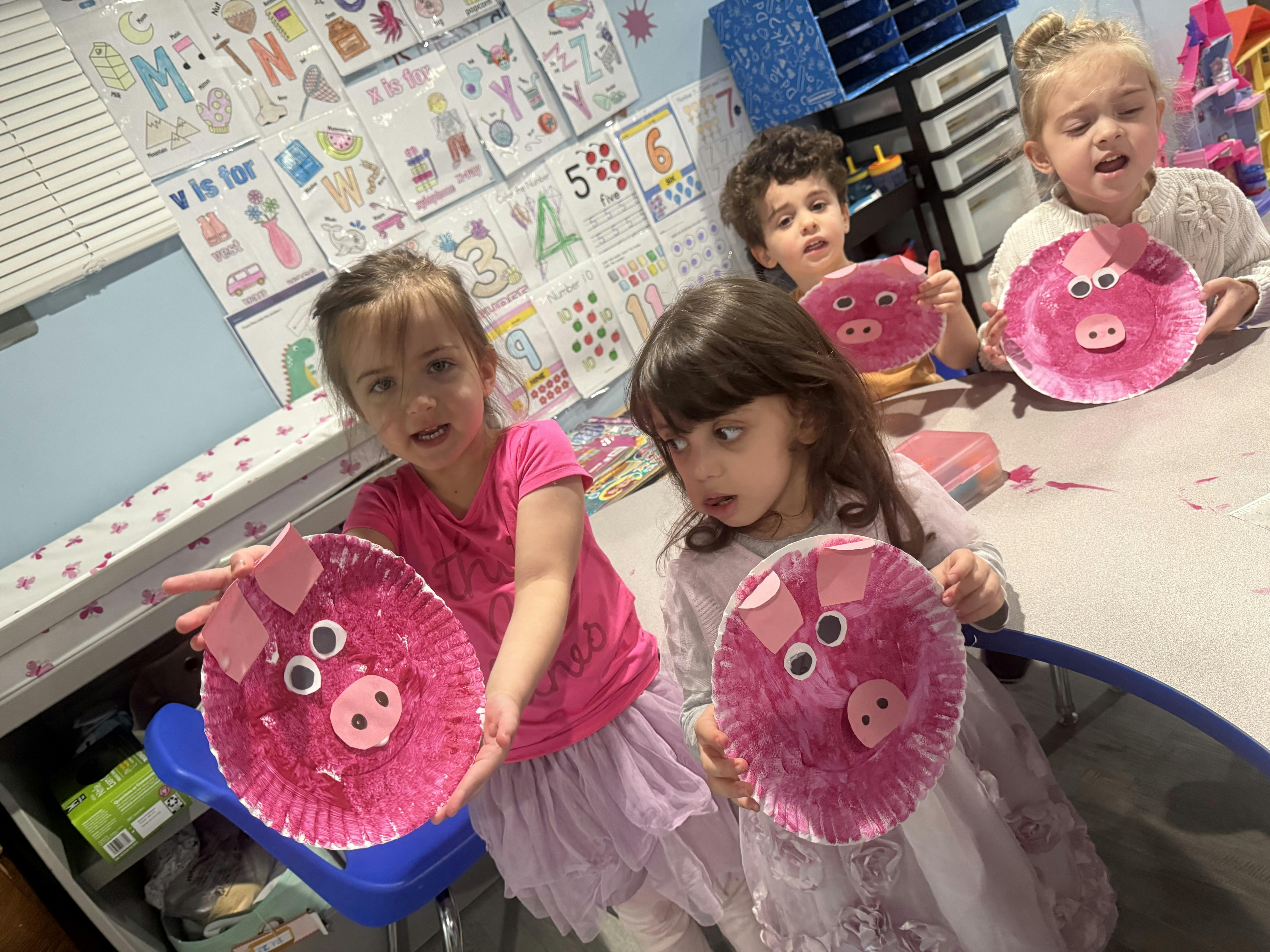Four children in a classroom holding up painted paper plate pigs