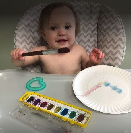 Child in high chair holding a paintbrush with watercolors and paper plate