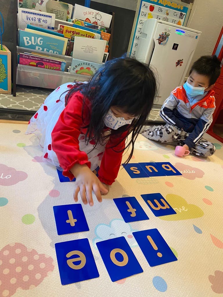 Child playing with letter cards on the floor, another child in background