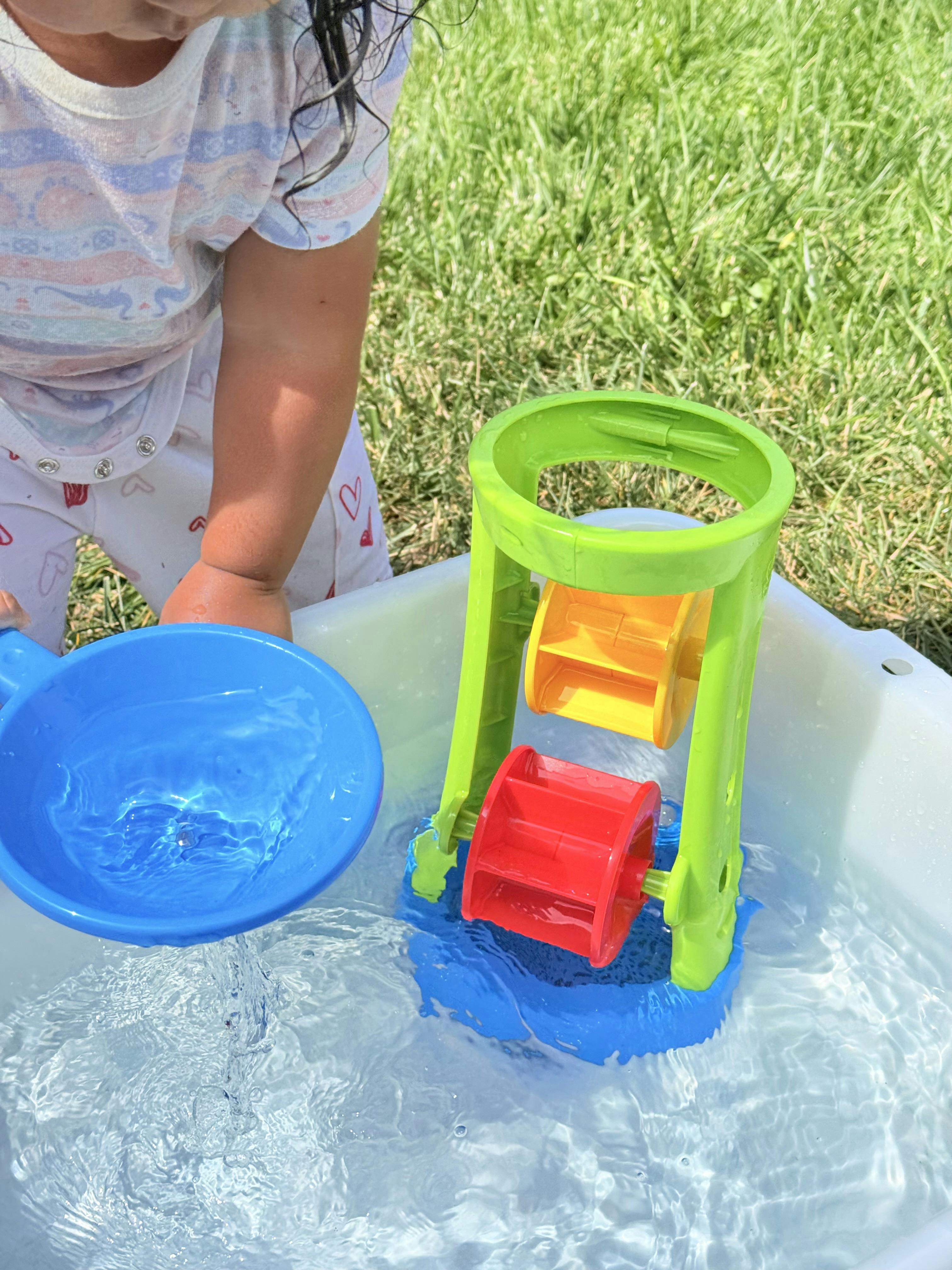 Child pouring water from a blue cup onto a toy water wheel in a bin