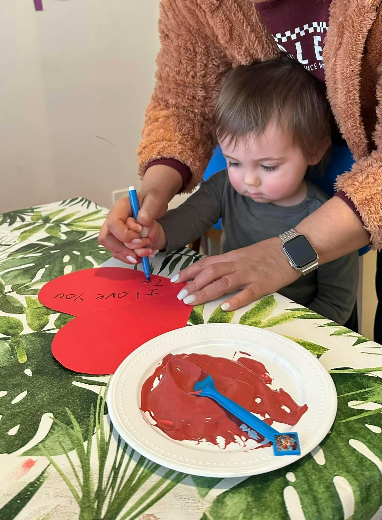 Person helping a child write on a red heart-shaped paper