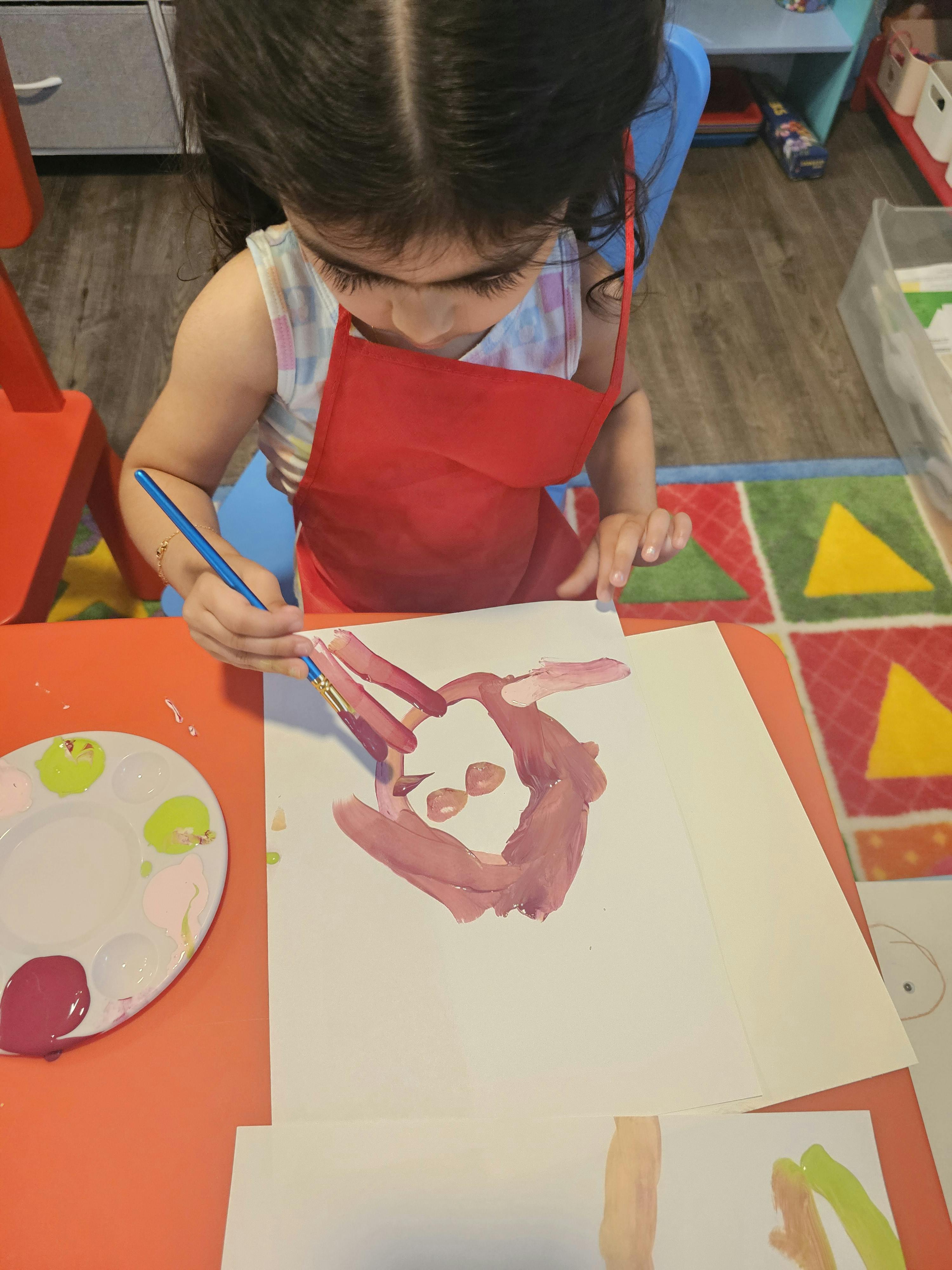 Child in a red apron painting on paper with a paintbrush