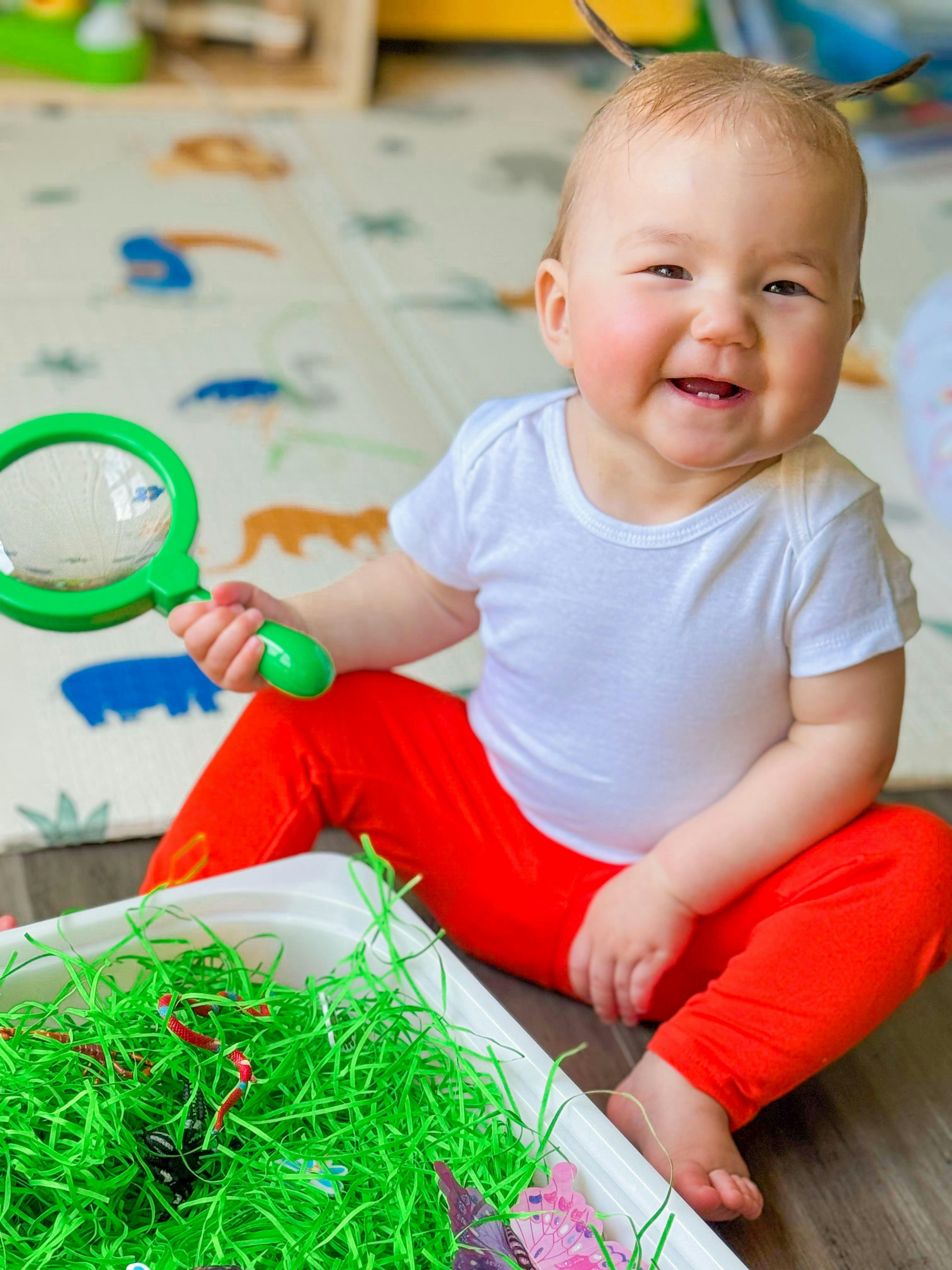 A child sitting on the floor holding a magnifying glass near a bin of grass