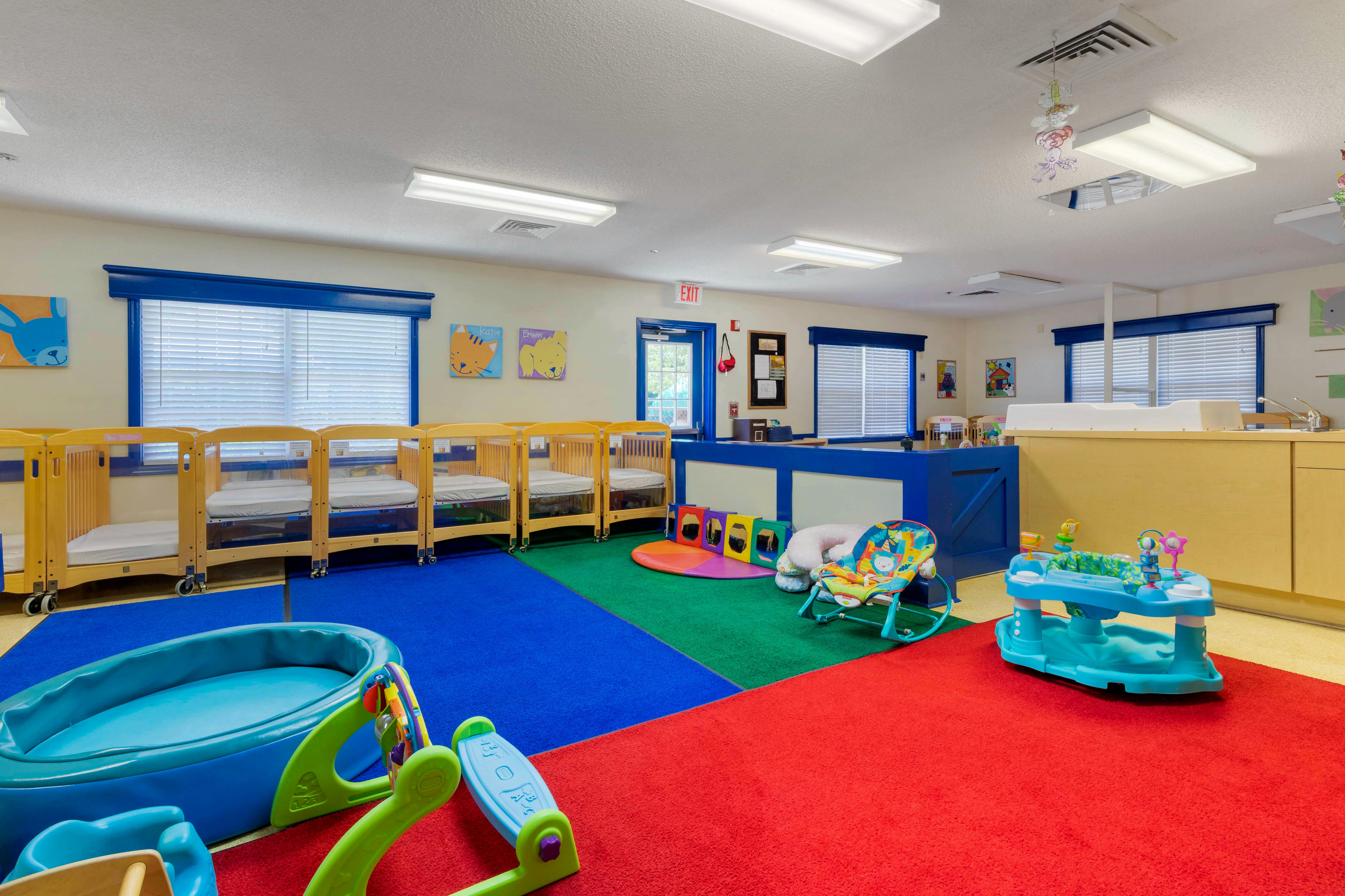 A colorful childcare room with cribs, play mats, and baby bouncers