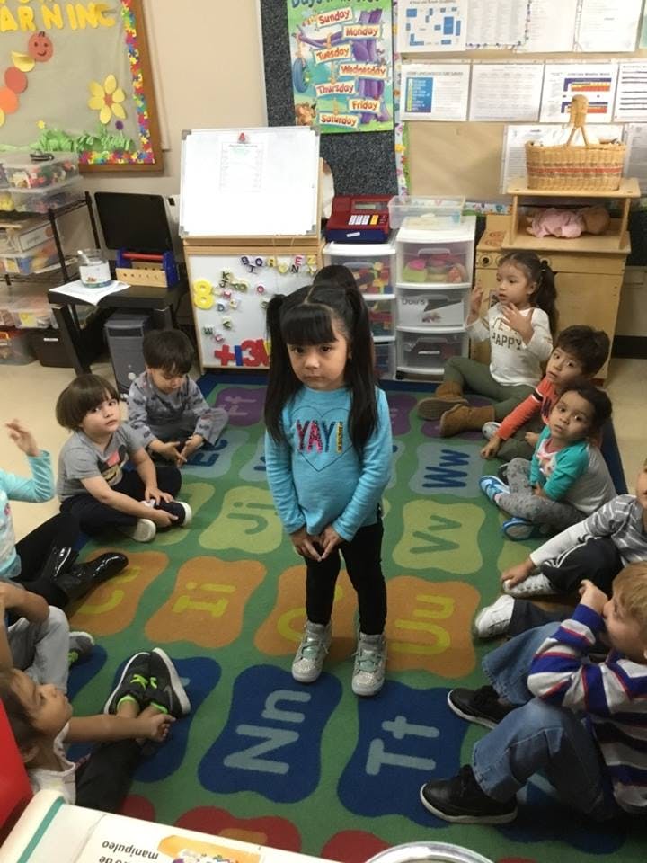 Child standing in a classroom with other children sitting on a rug
