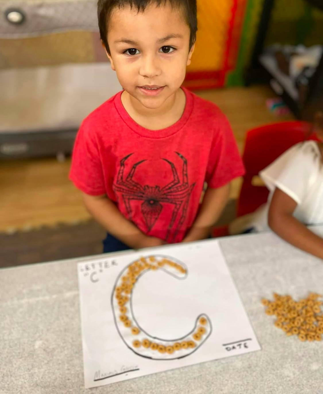 Child in red shirt with a craft of the letter C made from cereal