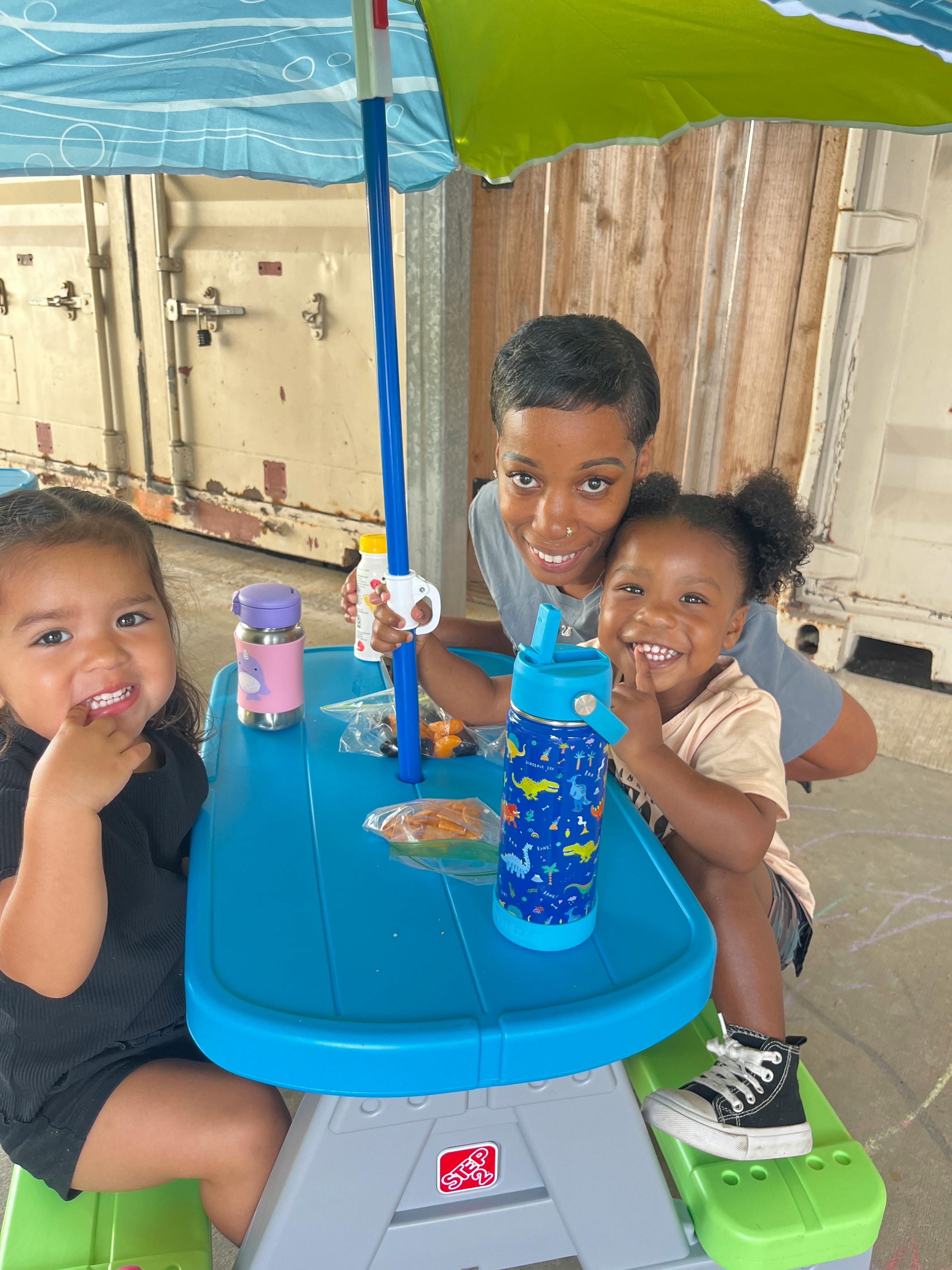 Person and two children sitting at a small blue picnic table with an umbrella