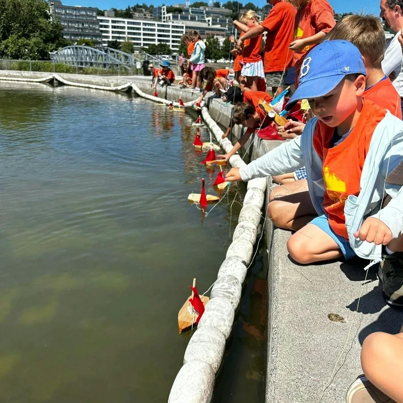 Child in blue hat and orange shirt reaching for small wooden boat in water