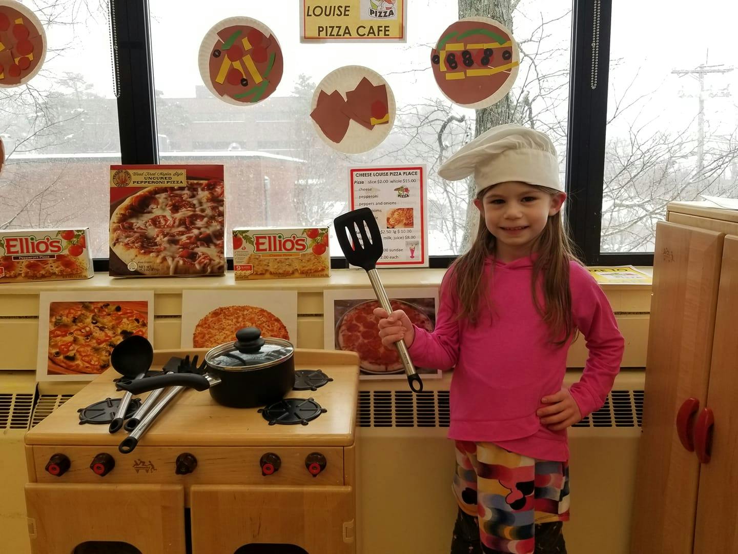 Child wearing a chef hat and holding a spatula next to a play kitchen