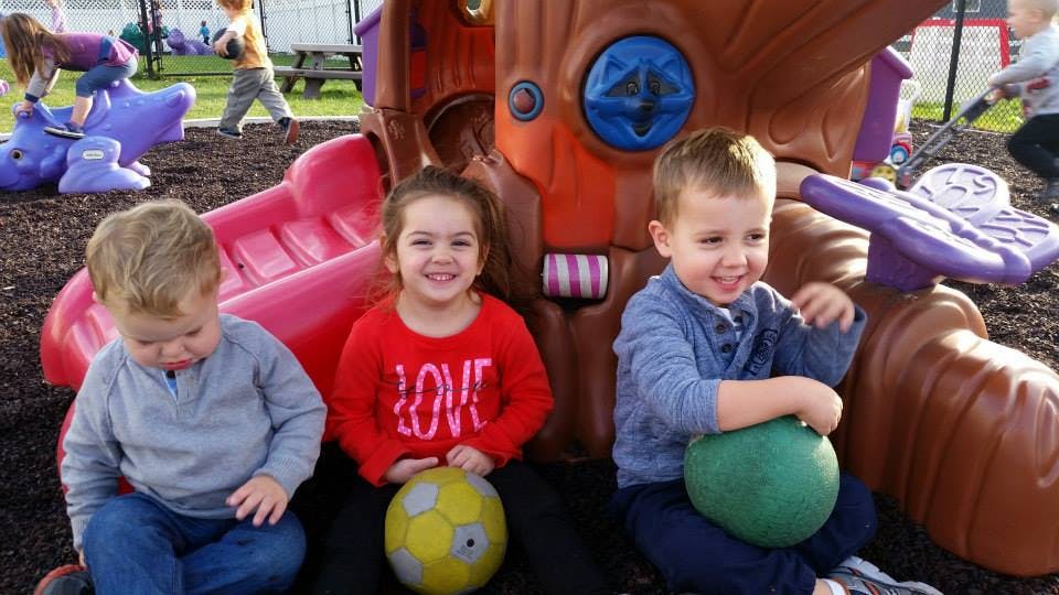Three children sitting on the ground with balls near a playground structure