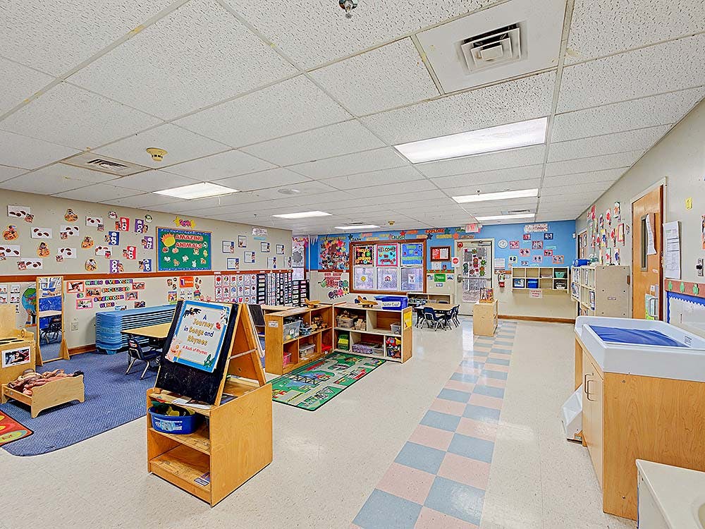 Empty classroom with small tables and chairs, shelves with toys, and decorated walls