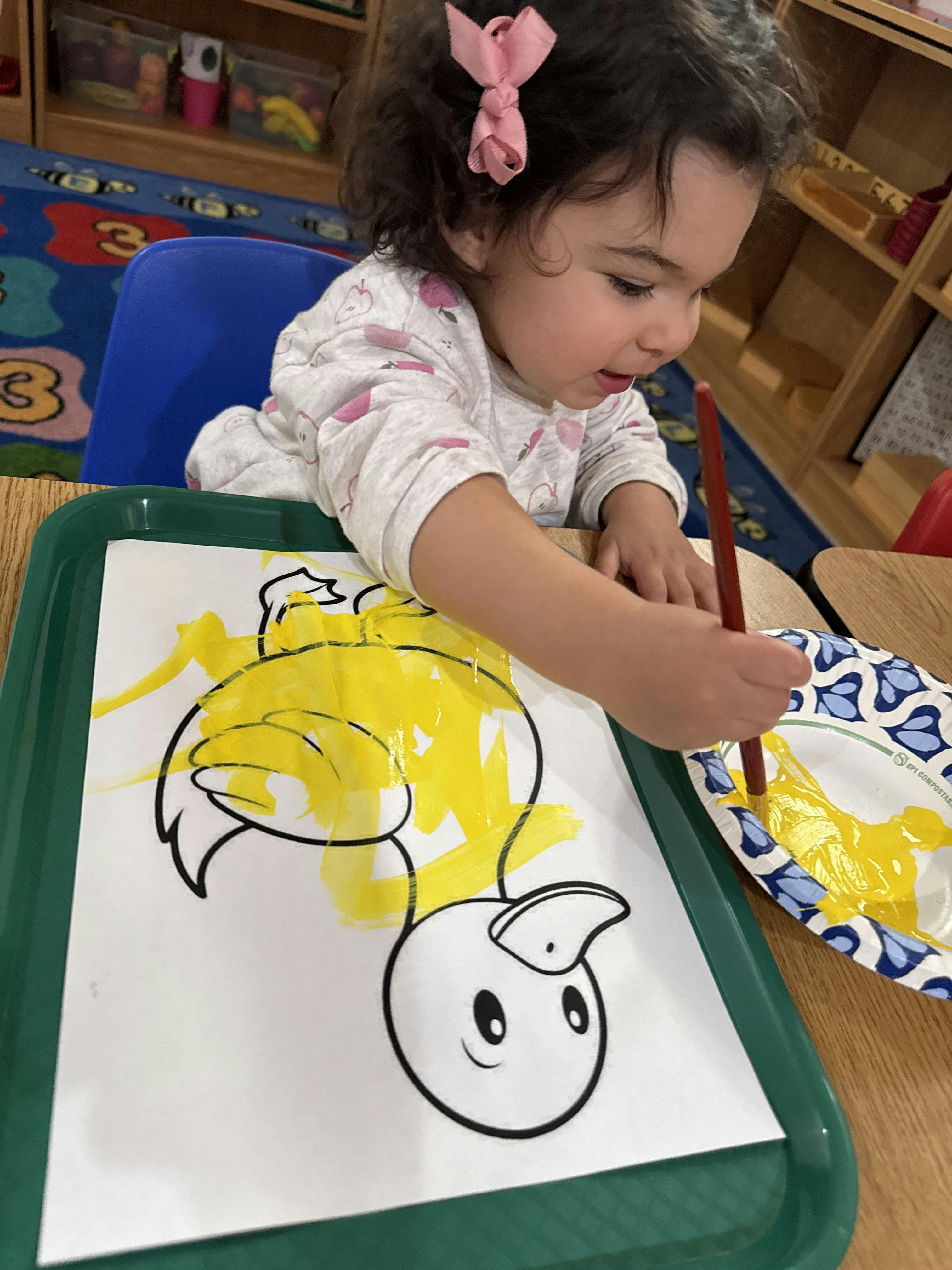 Child coloring a duck illustration with yellow paint in a classroom