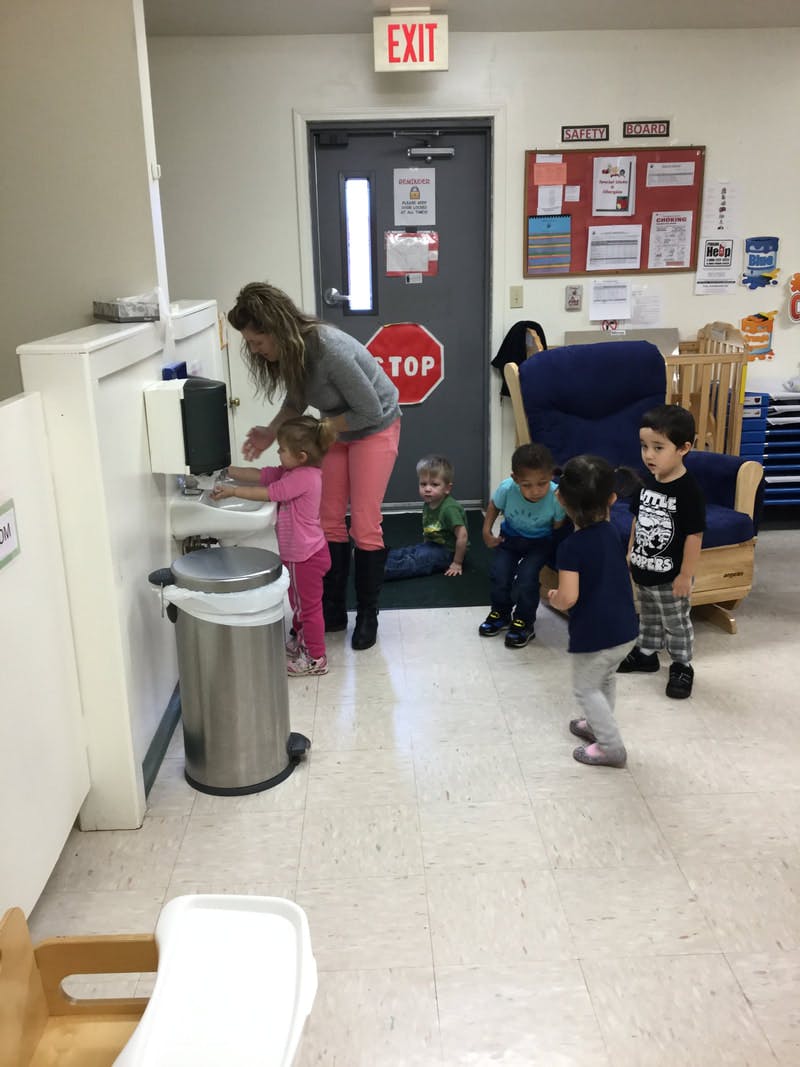 Adult helping a child wash hands at a sink, other children nearby