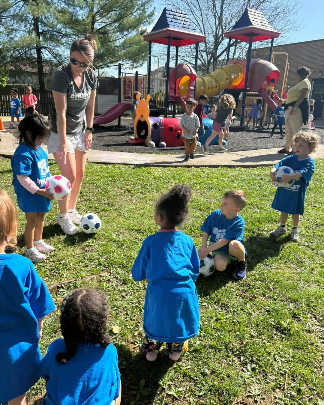 Adult supervising children with soccer balls on grass next to a playground