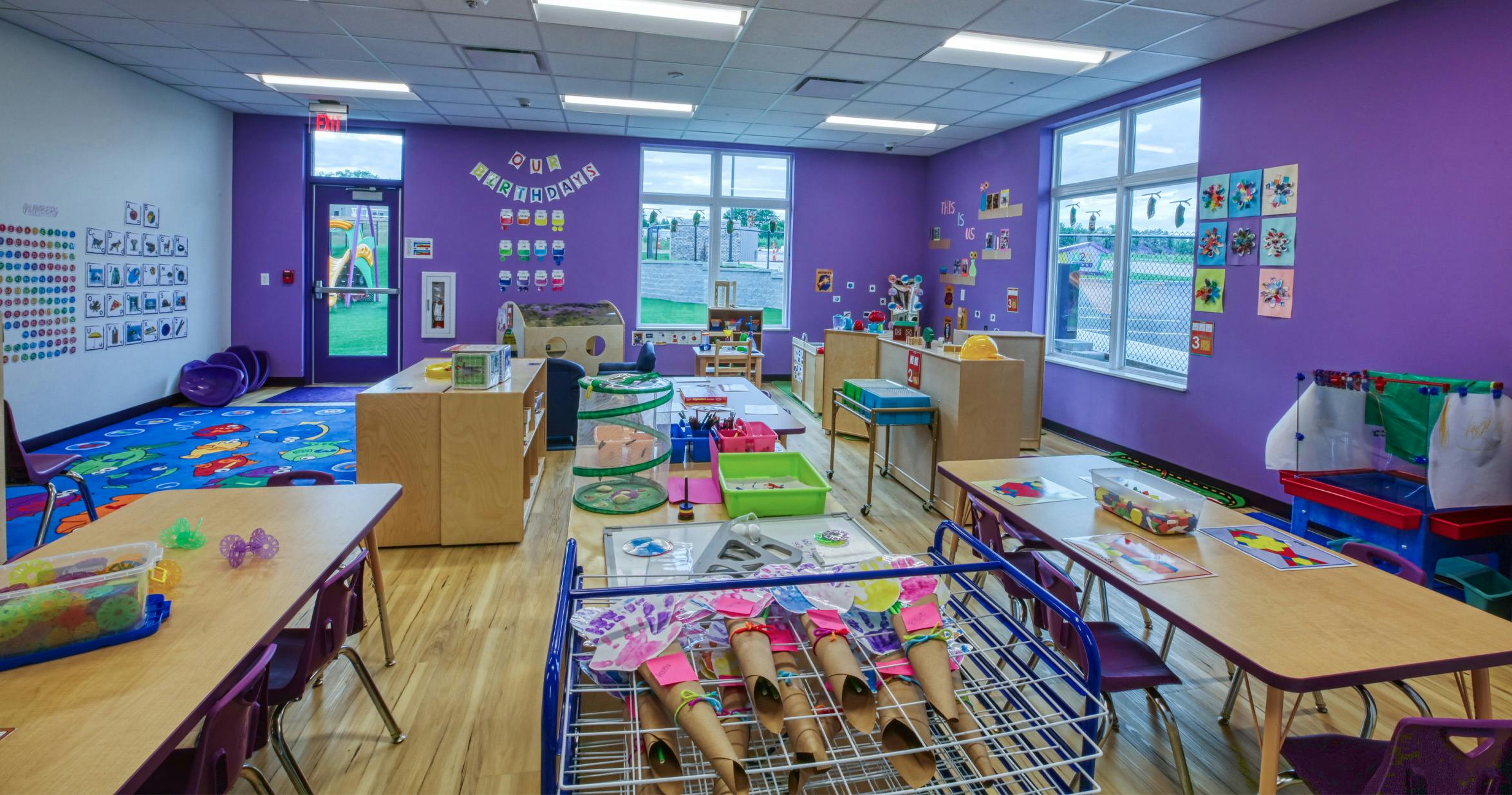 Colorful classroom with tables, chairs, learning materials, and playground visible outside
