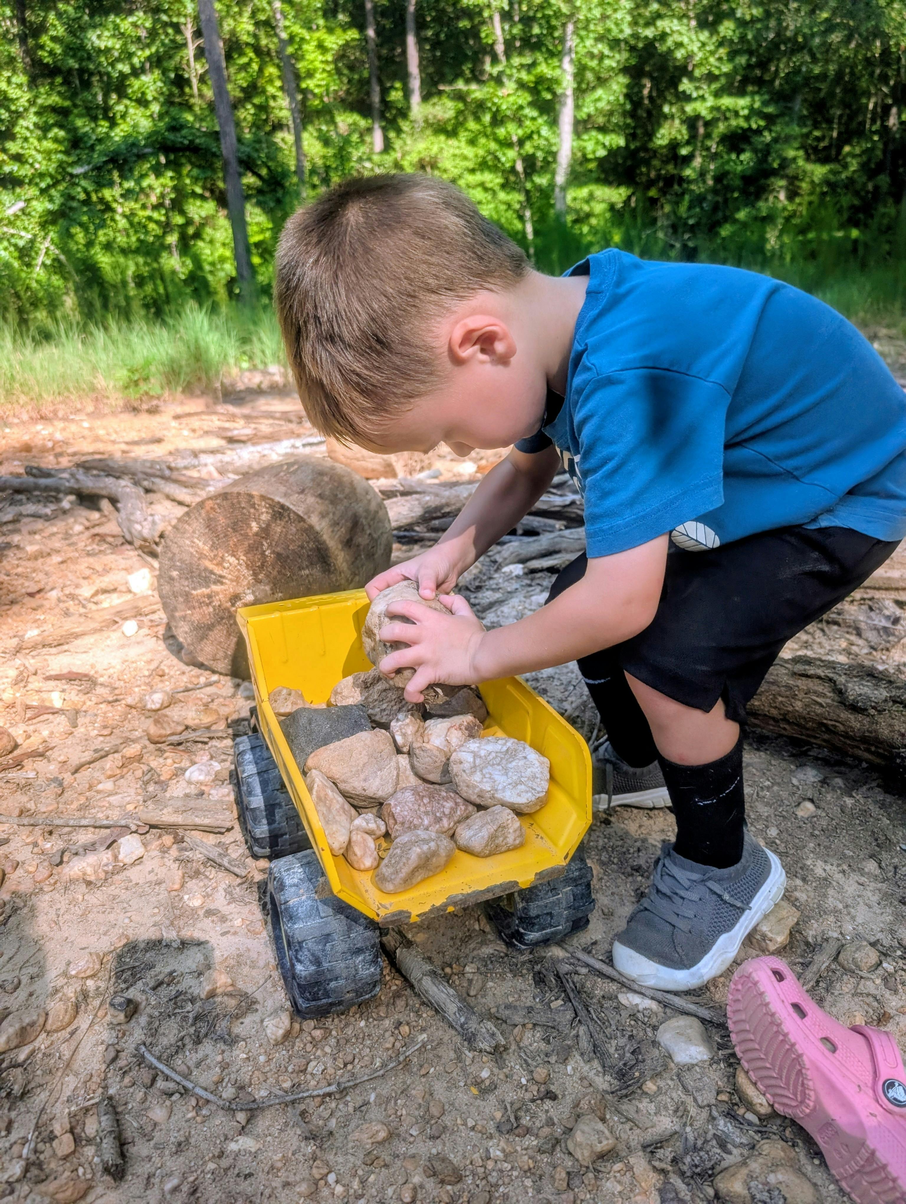 Child placing rocks into a toy dump truck in a wooded area