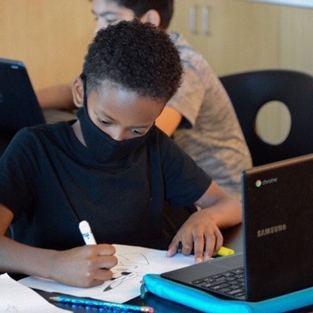 Child in a mask drawing at a desk with a laptop