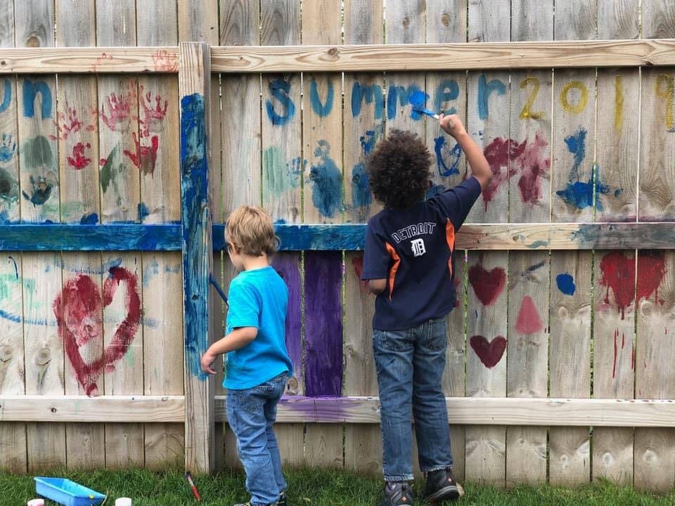 Two children painting on a wooden fence with colorful paints