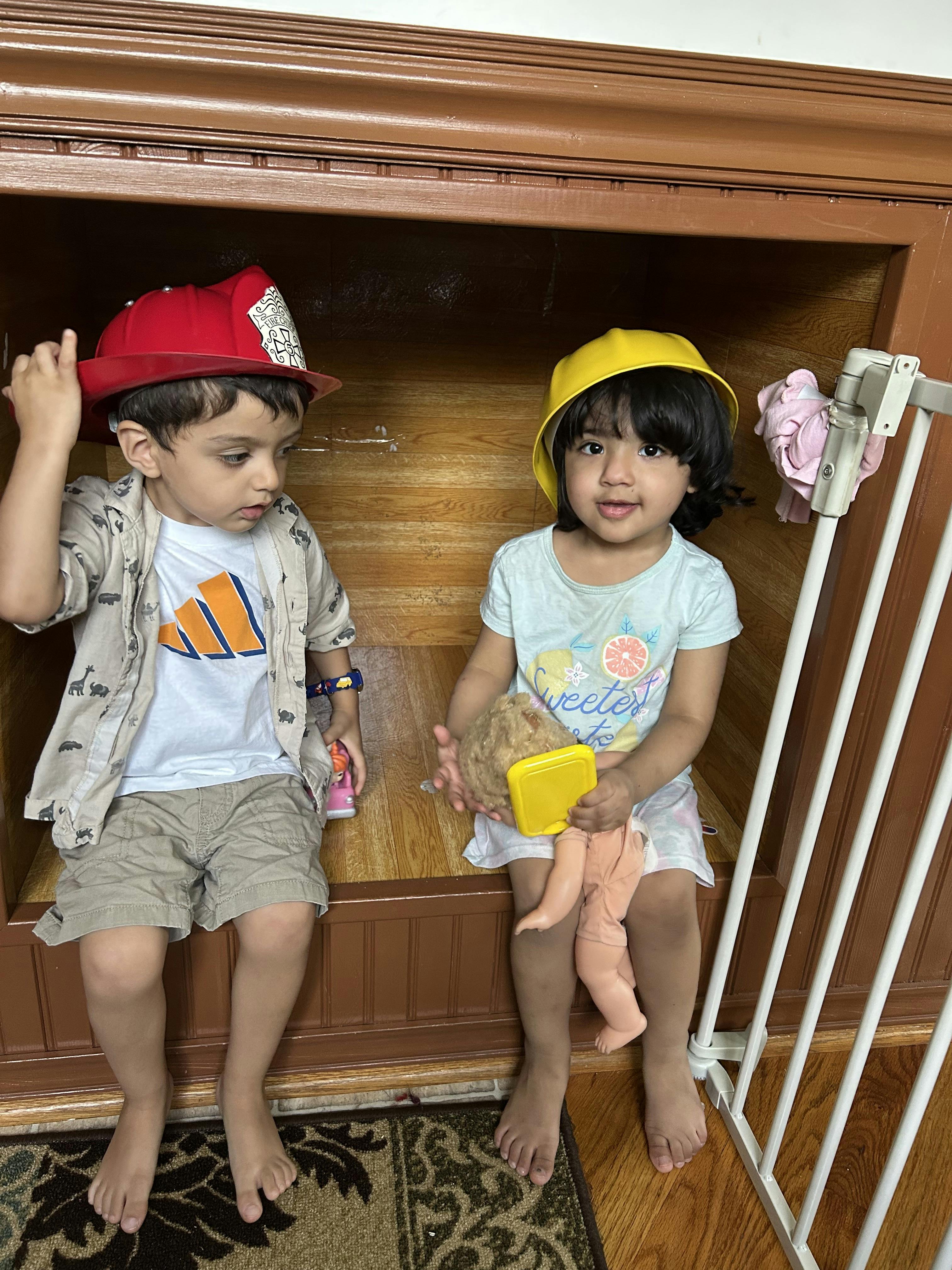Child in a red firefighter hat and a child in a yellow hard hat holding a doll