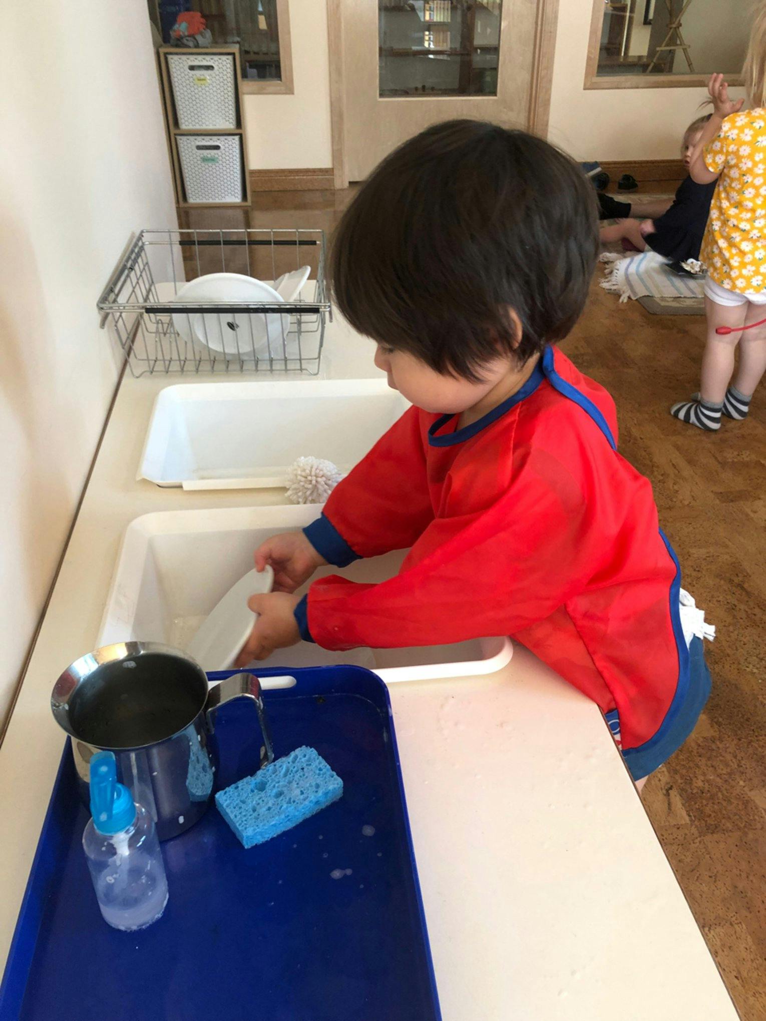 Child in a red smock washing dishes in a sink