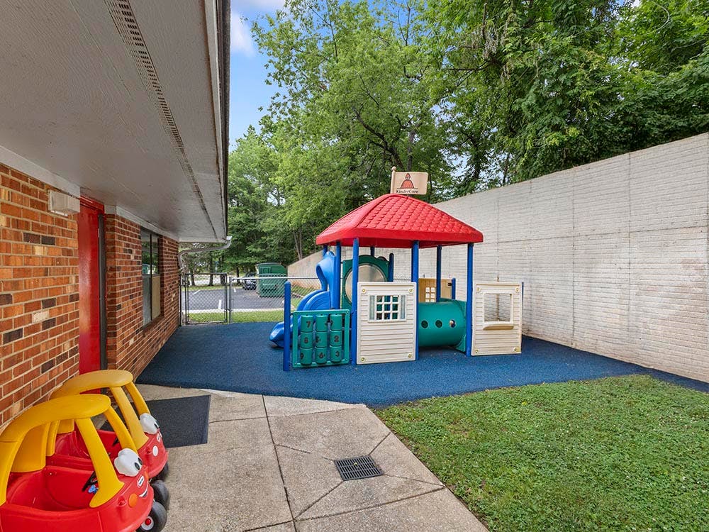 Playground with blue padded ground next to a brick building, two red toy cars