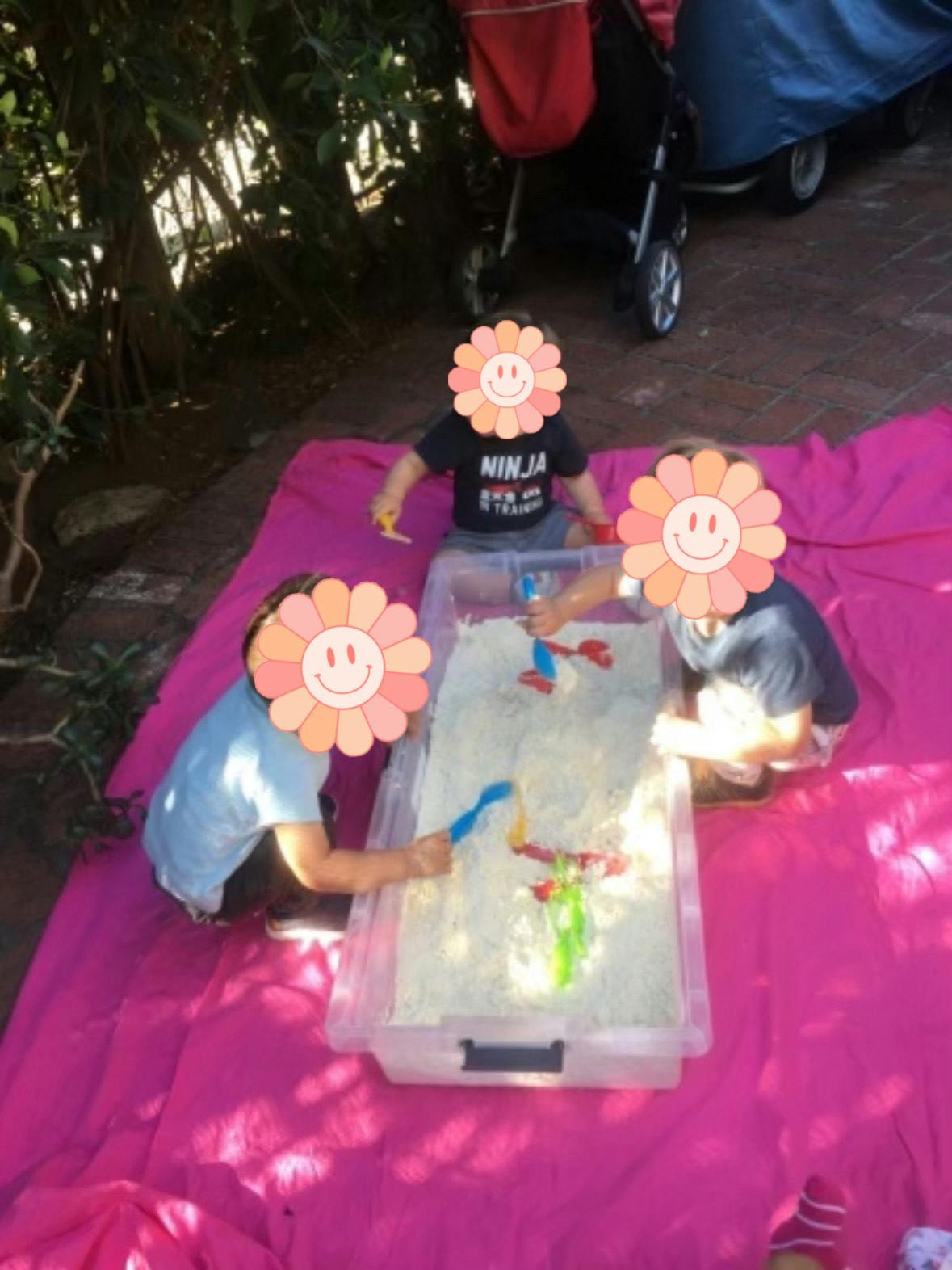 Three children playing with sand in a container on a pink blanket