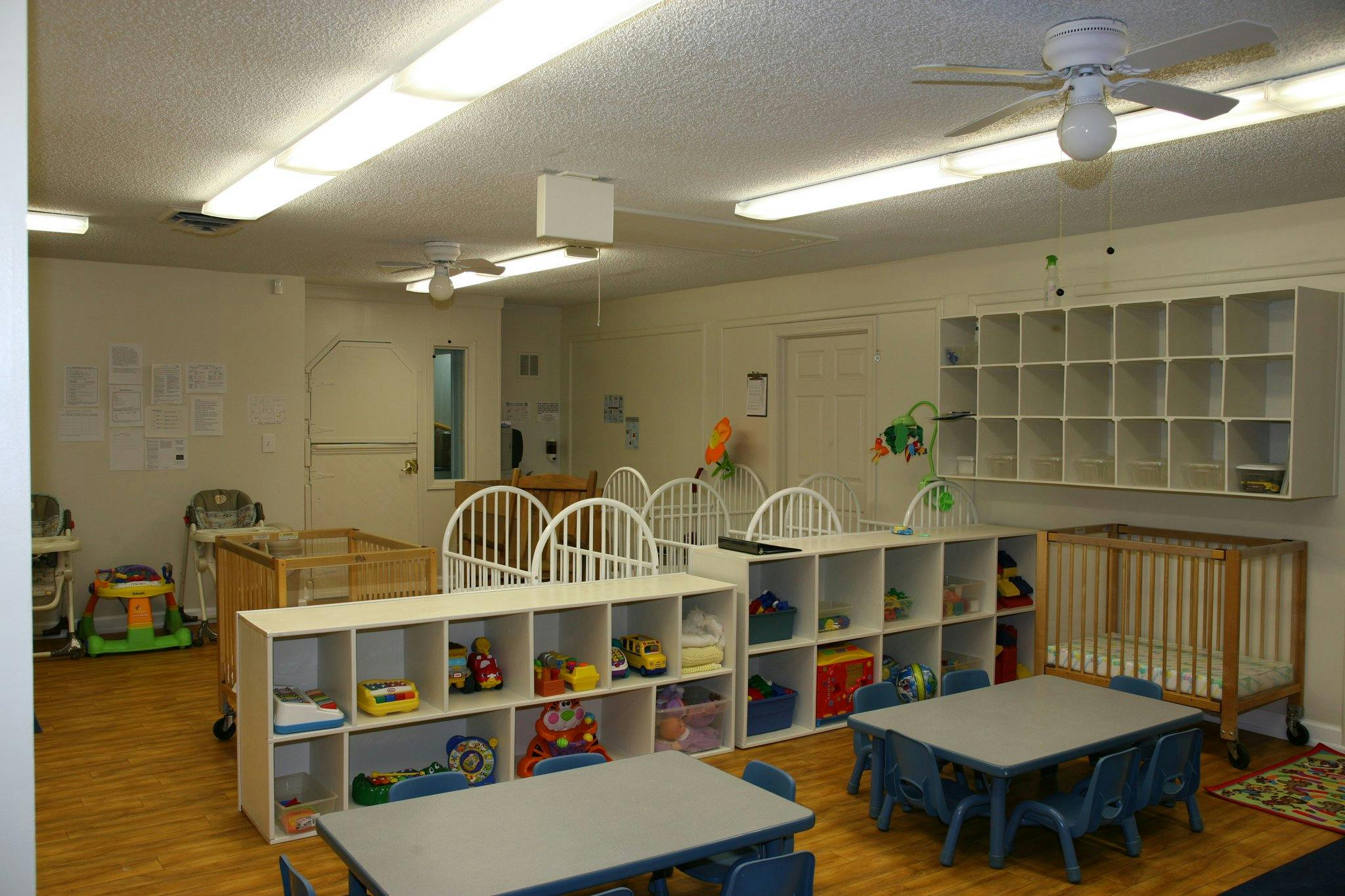 Daycare room with cribs, shelves of toys, and small tables with chairs
