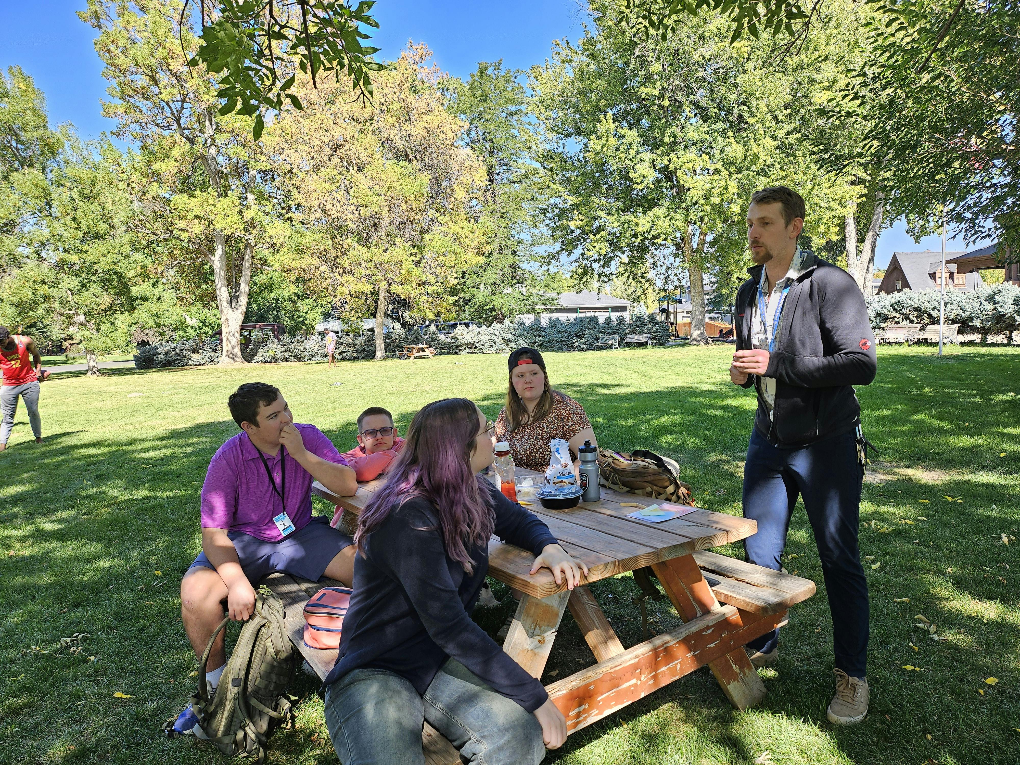 People gathered around a picnic table outdoors, one person standing and talking