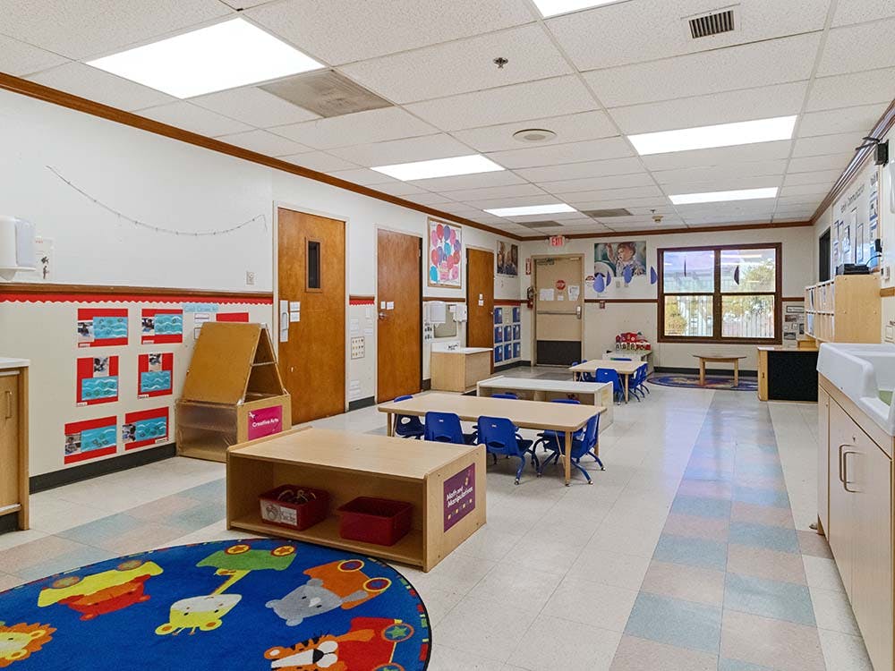 Empty classroom with small tables and chairs, a play rug, and activity centers