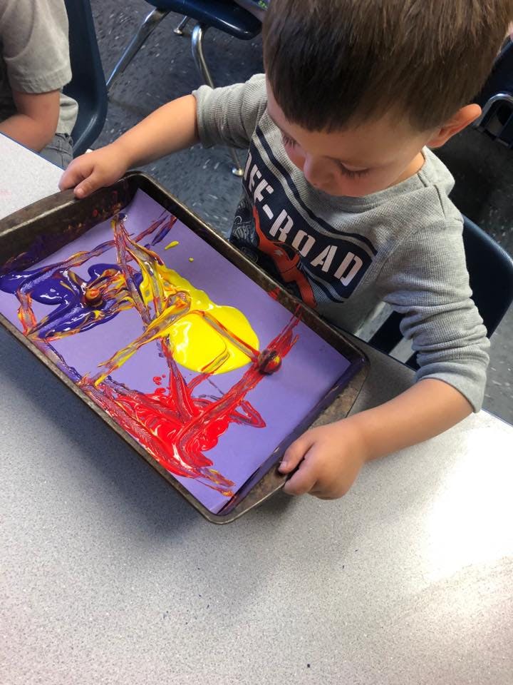 Child holding a tray with a purple paper covered in red, yellow, and purple paint