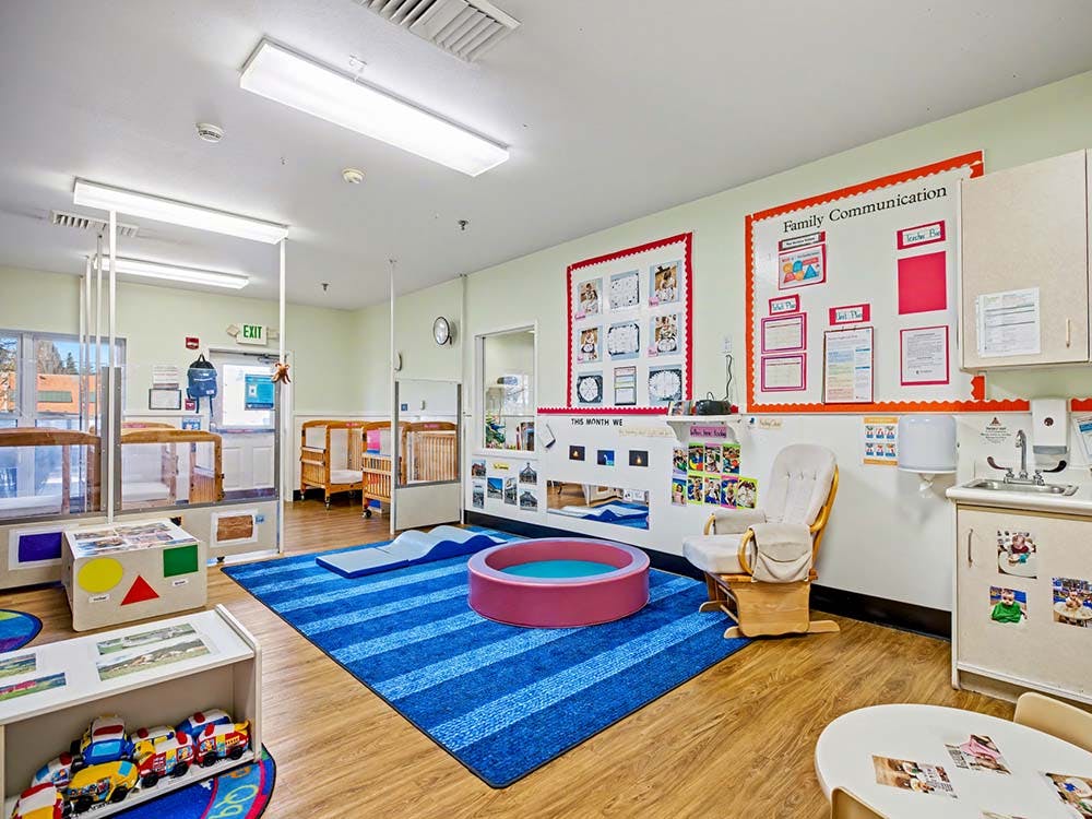 A colorful childcare classroom with cribs, play mats, and a small sink area